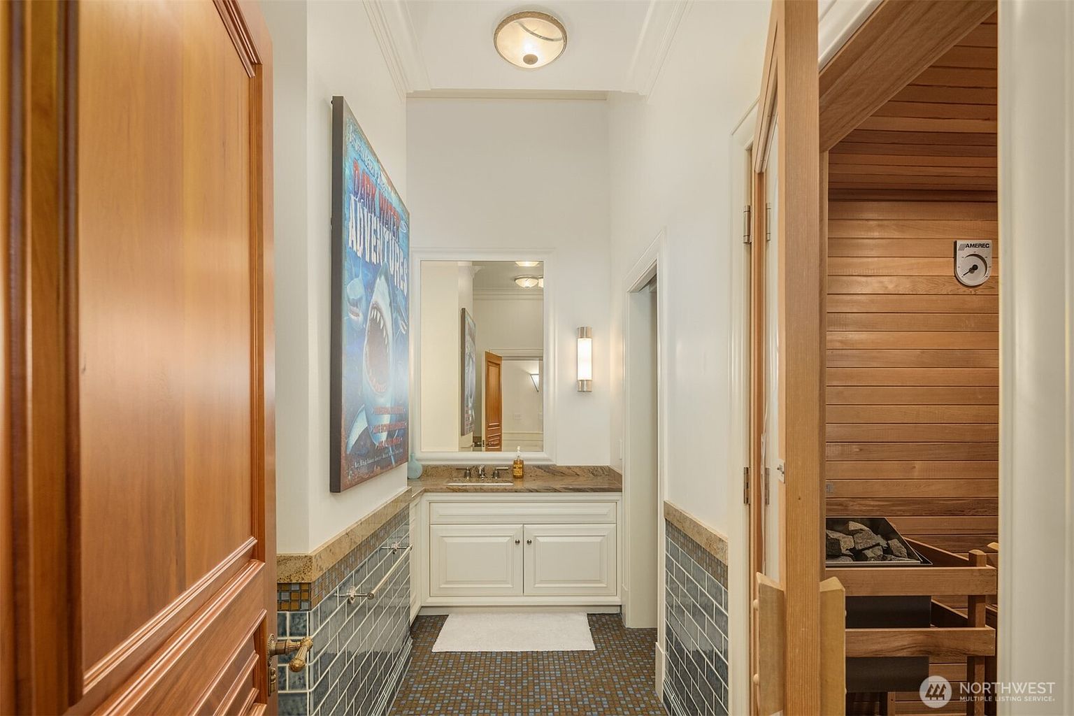 This interior view captures a unique bathroom space featuring a built-in sauna on the right with wood-paneled walls and a stone heater. The vanity area includes a white cabinet with a granite countertop, a framed mirror, and a decorative poster on the wall, all set against a backdrop of blue-tiled wainscoting and dark floor tiles. The perspective is from a doorway, looking into the functional and spa-like room.