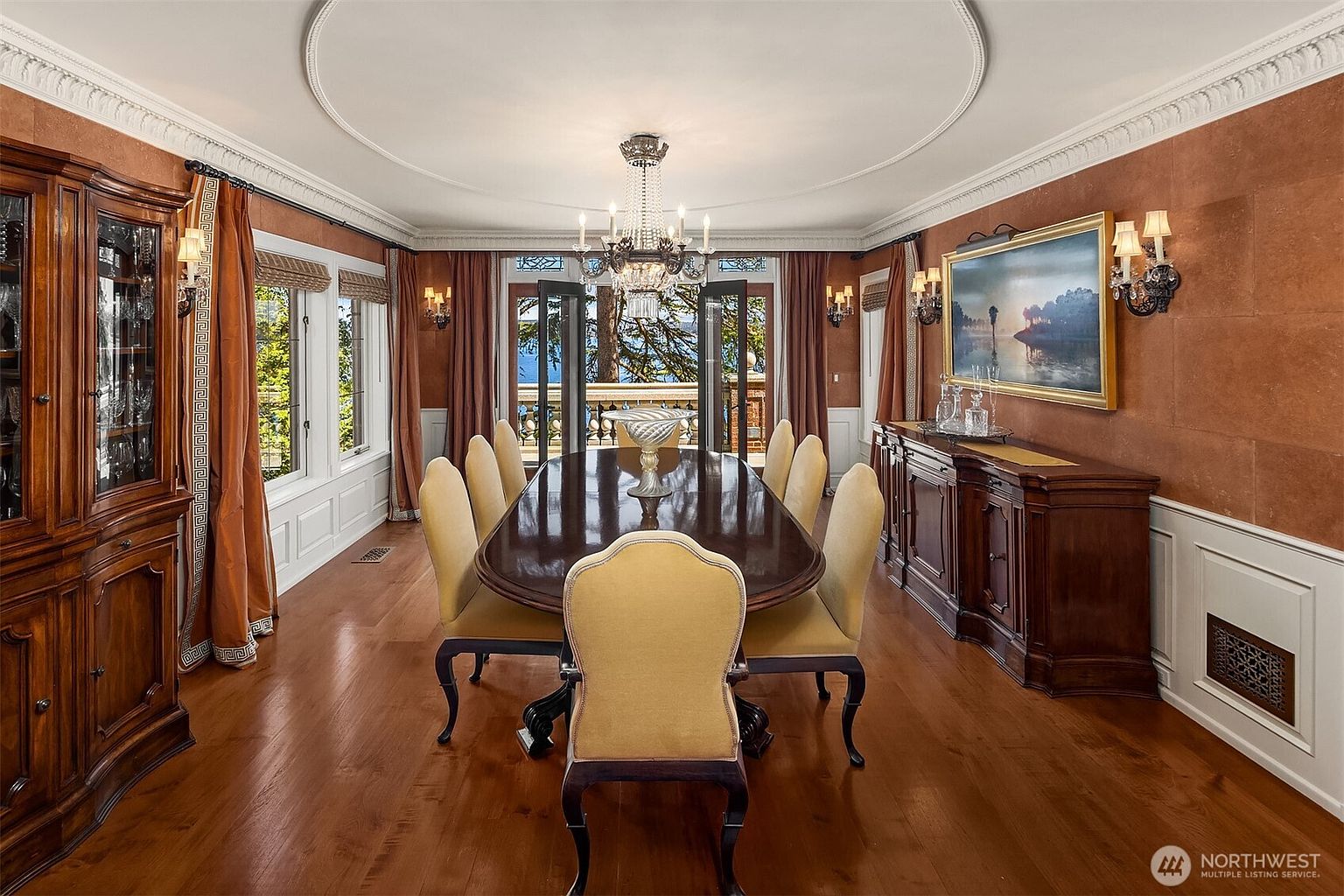 This elegant formal dining room features a rich, traditional aesthetic with warm, textured terracotta-colored walls and polished hardwood floors. A grand, dark wood dining table is surrounded by upholstered chairs, centered beneath a sophisticated crystal chandelier that hangs from a detailed ceiling medallion. The room is framed by large windows and French doors that offer a scenic view of the outdoors, complemented by a matching wooden hutch and sideboard.