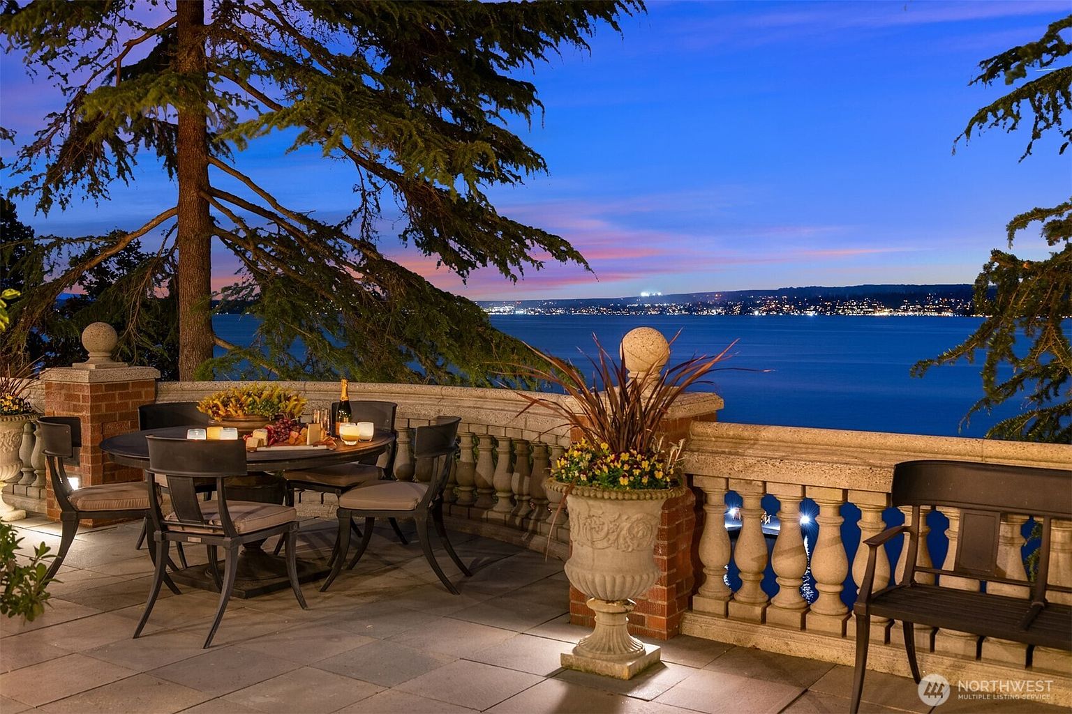 This elegant stone terrace offers a breathtaking, elevated view of a tranquil lake at twilight, framed by a classic balustrade and a large, mature tree. The space is set up for intimate outdoor dining with a round table, comfortable chairs, and a decorative stone planter, all illuminated by soft, warm accent lighting. The cinematic perspective captures the serene transition from the cozy, well-appointed patio to the expansive, glowing cityscape across the water.