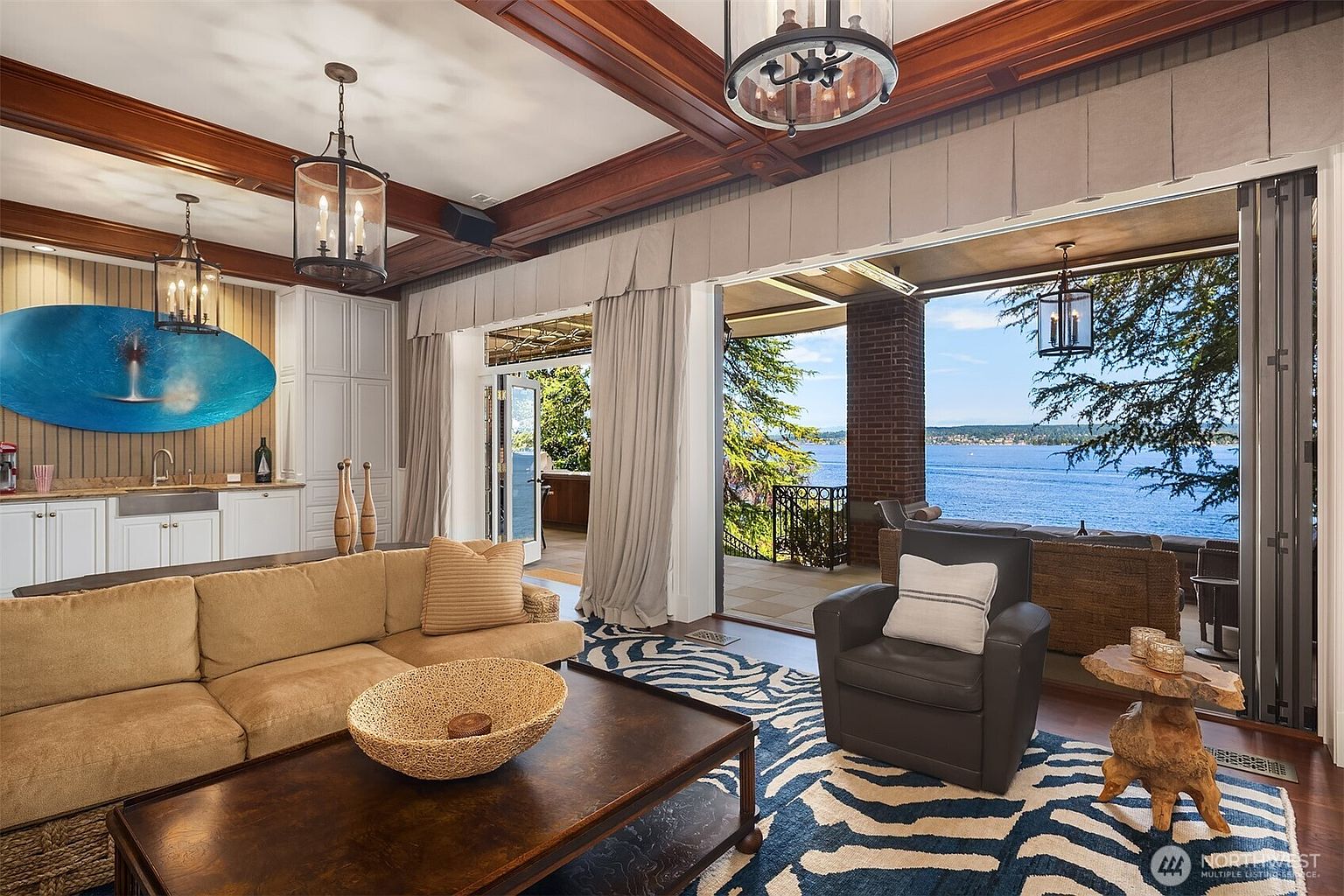 This elegant living room features a warm, inviting color palette with a plush tan sectional sofa and a striking blue and white zebra-print area rug. The space seamlessly transitions to a covered outdoor patio through large, open glass doors, offering a breathtaking, unobstructed view of the lake. Architectural details like the coffered wood ceiling and classic lantern-style chandeliers add a touch of sophisticated, traditional charm to the open-concept design.