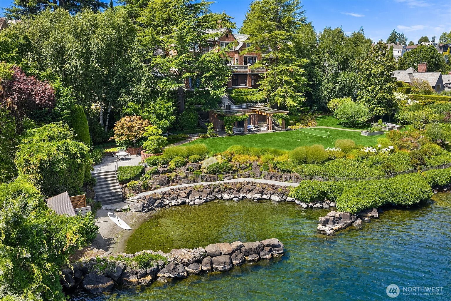 This high-angle aerial view captures a grand, multi-story brick estate nestled into a lush, terraced hillside overlooking a serene lake. The property features a meticulously manicured lawn, a covered patio area, and a private stone-walled beach access point with a paddleboard resting on the shore. The scene exudes luxury and tranquility, showcasing the seamless integration of classic architecture with expansive, nature-focused outdoor living spaces.