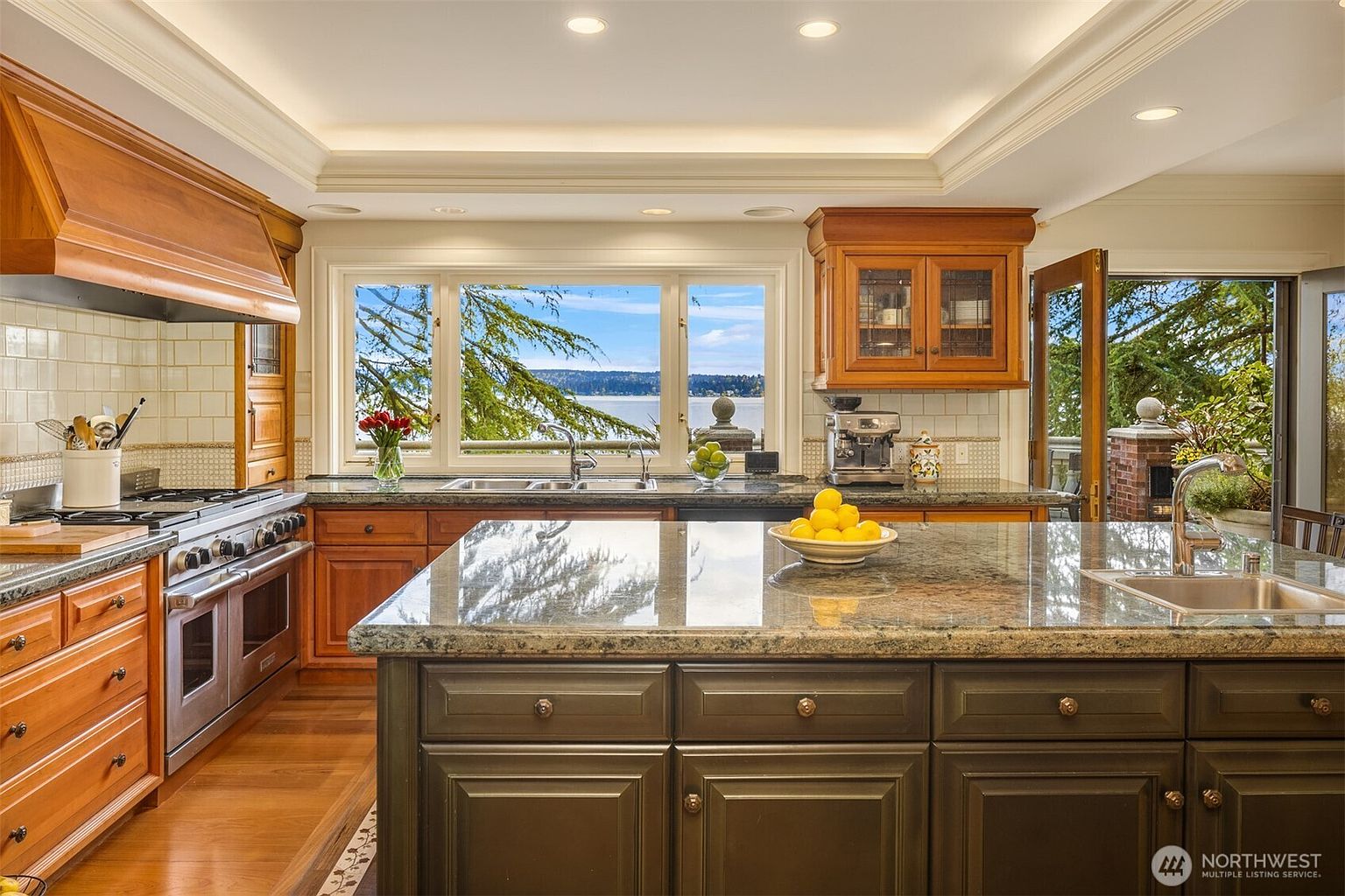 This elegant, traditional-style kitchen features warm wood cabinetry, granite countertops, and a large central island with a dark green base. A spacious window above the sink provides a stunning, unobstructed view of the water and trees, while high-end stainless steel appliances and a professional-grade range hood add a touch of luxury. The space is well-lit with recessed lighting, creating a bright and inviting atmosphere perfect for both cooking and entertaining.