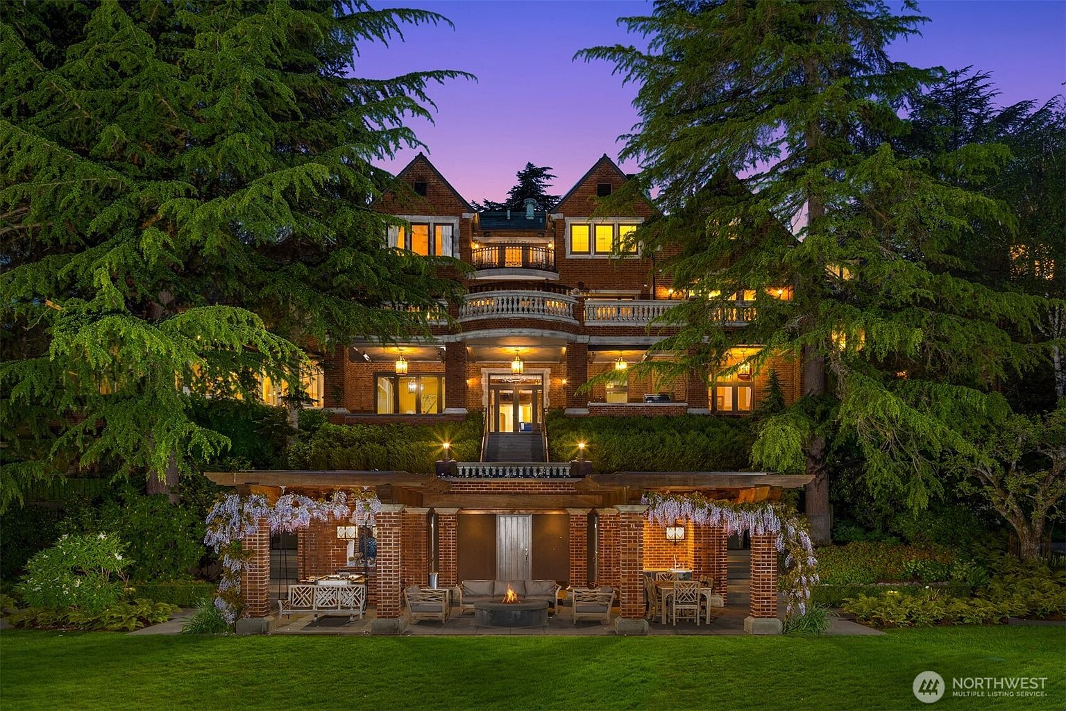 This grand, multi-story brick estate is captured at twilight, showcasing its impressive architectural scale and tiered landscaping. The foreground features a charming, wisteria-draped pergola with a cozy outdoor seating area and fire pit, leading up a central staircase to the main residence. The warm glow from the interior windows and exterior lighting creates an inviting and luxurious atmosphere against the deep purple dusk sky.