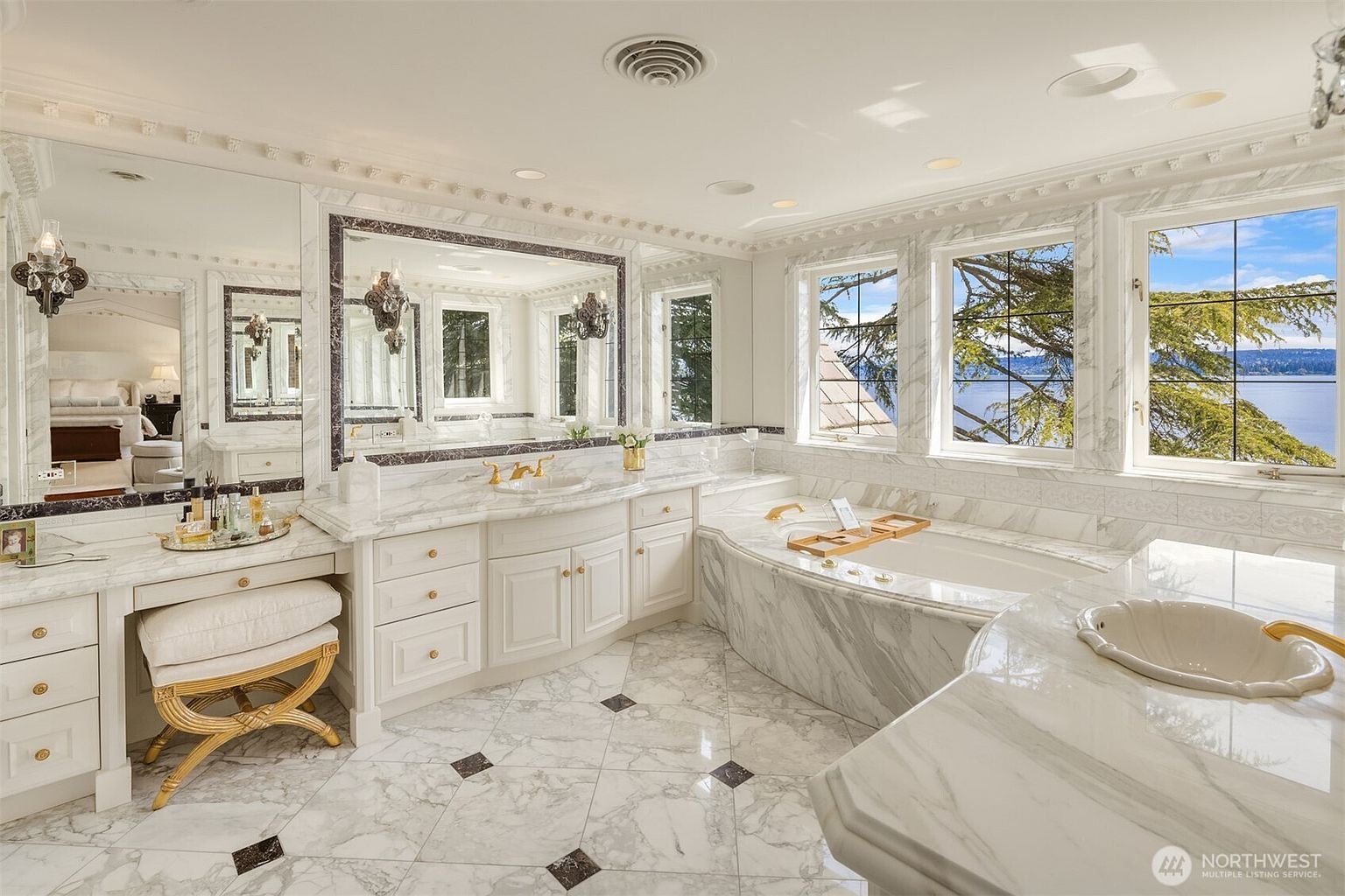 This luxurious primary bathroom features elegant white marble countertops and flooring with dark accent tiles, creating a sophisticated and timeless aesthetic. The space includes a spacious soaking tub positioned by large windows that offer a stunning, panoramic view of the water, alongside a dedicated vanity area with a gold-accented stool. The room is bright and airy, reflecting a high-end, spa-like atmosphere perfect for relaxation.