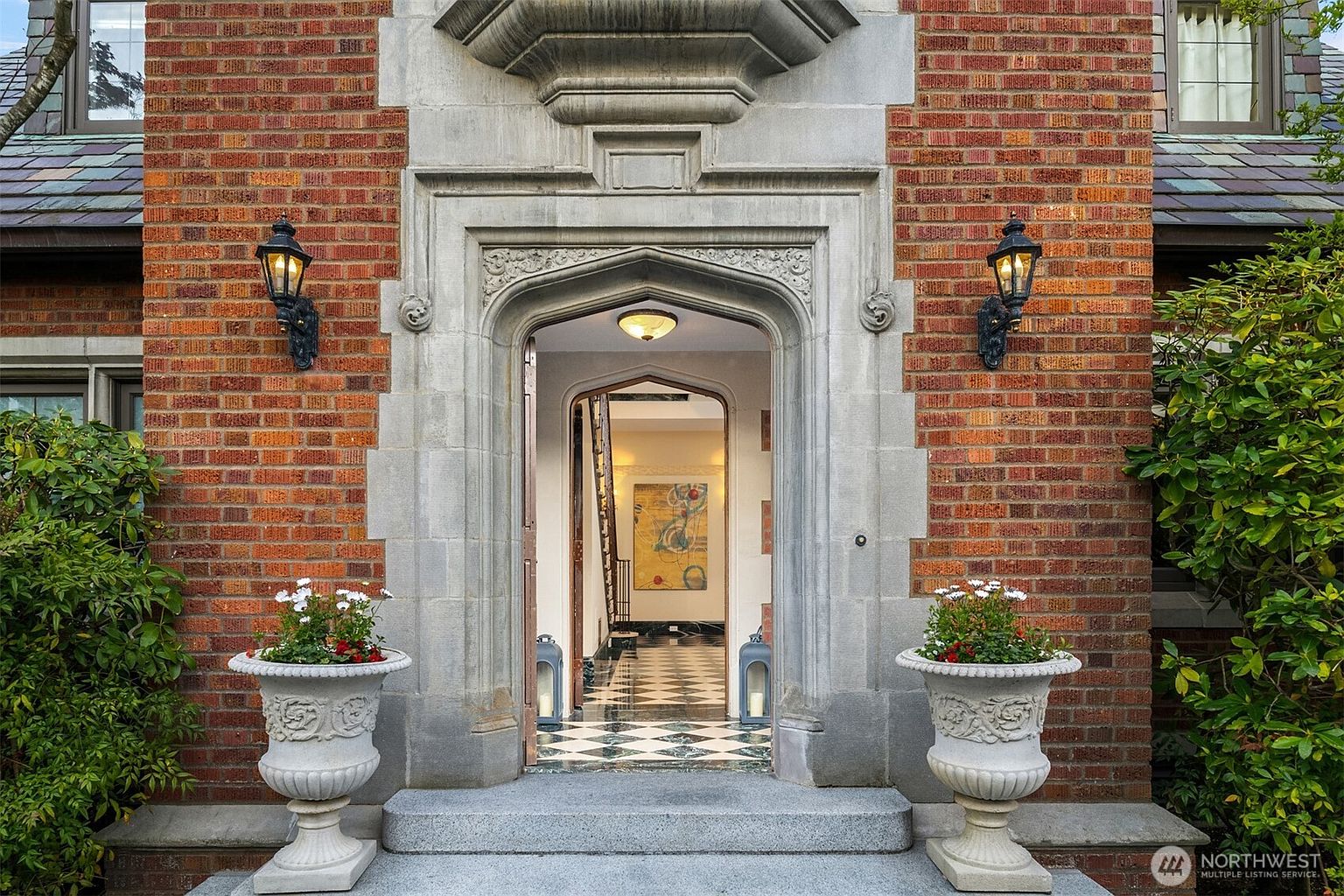 This elegant front entryway features a classic Tudor-style brick facade with a prominent, ornate stone-carved archway framing the front door. Two large, decorative stone urns filled with flowers flank the entrance, while traditional wall-mounted lanterns provide a warm, inviting glow. The perspective looks directly into the foyer, revealing a sophisticated black-and-white checkered marble floor and a glimpse of an interior art piece.
