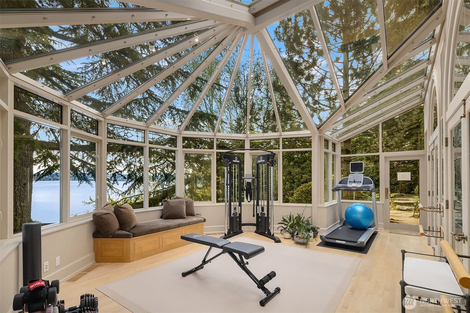 This bright and airy sunroom has been converted into a home gym, featuring a glass-enclosed conservatory design that offers panoramic views of the surrounding trees and water. The space is equipped with a functional cable machine, a weight bench, and a treadmill, all set upon a neutral area rug. The combination of natural light, high-end fitness equipment, and serene outdoor scenery creates an inviting and motivating workout environment.