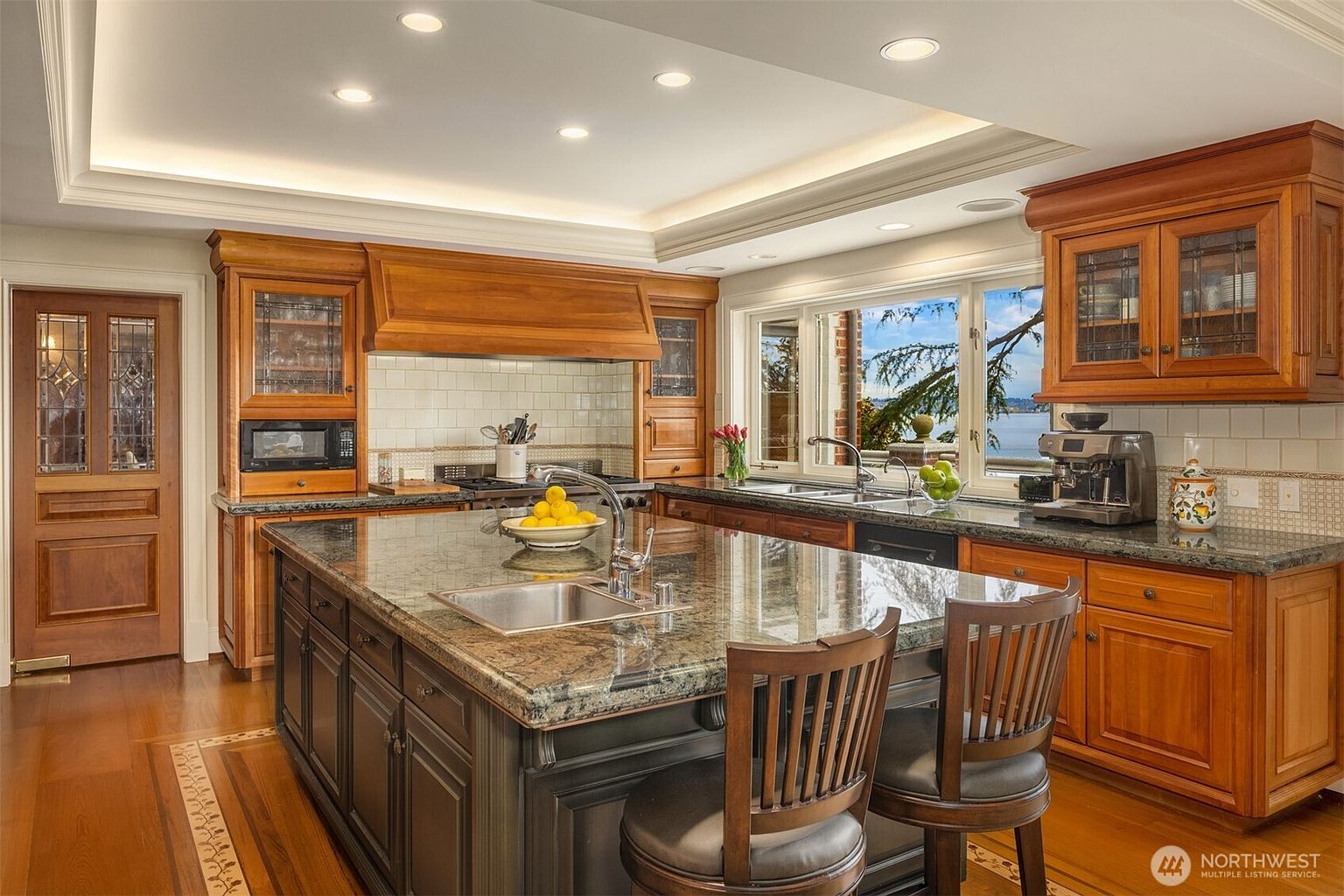 This elegant, traditional-style kitchen features rich wood cabinetry, a large central island with a dark granite countertop, and a built-in sink. The space is well-lit by recessed lighting and natural light streaming through a large window that offers a scenic view of the water. The combination of dark island cabinetry and warm wood tones creates a sophisticated and inviting atmosphere perfect for cooking and entertaining.