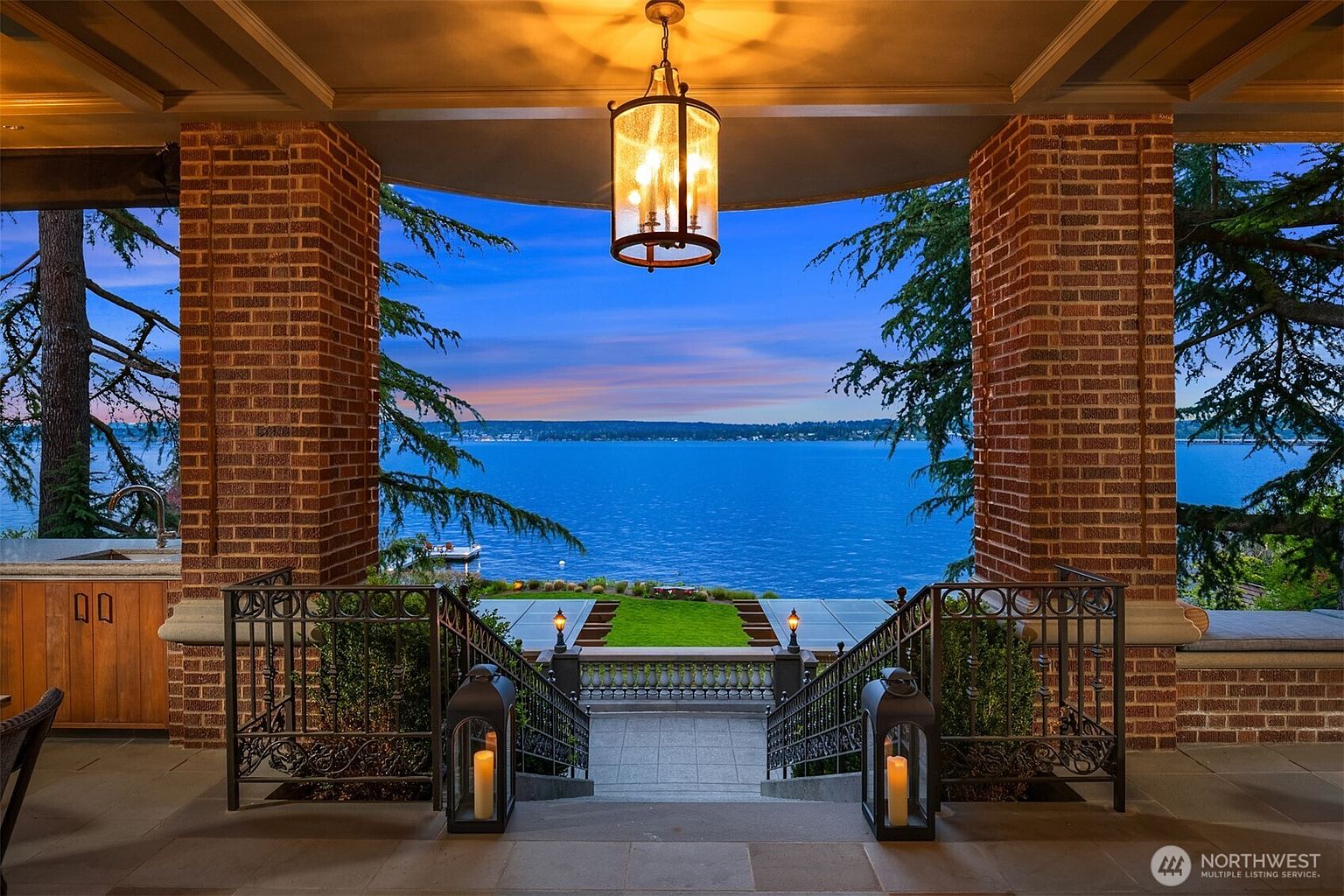 This elegant covered patio offers a breathtaking, unobstructed view of a serene lake at dusk, framed by sturdy brick columns. The space features a stone staircase leading down to a manicured lawn, accented by ornate wrought-iron railings and sophisticated lantern-style lighting. The combination of warm architectural textures and the expansive, tranquil water view creates a luxurious and inviting atmosphere.
