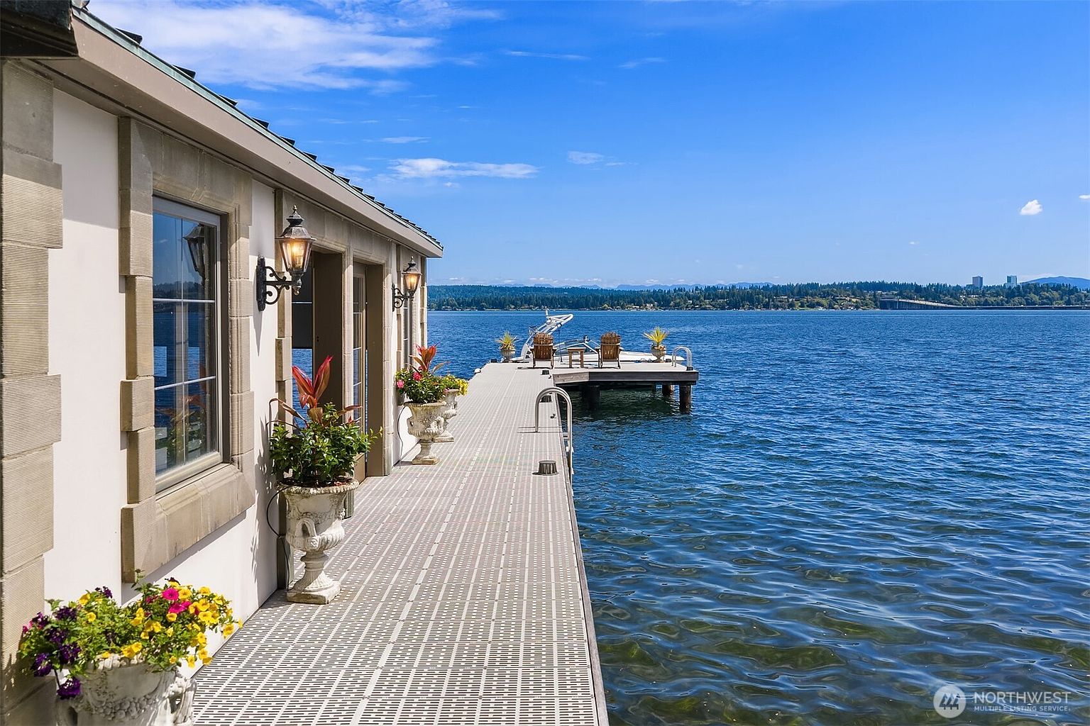 This image captures a serene waterfront deck attached to a home, extending directly over a calm, blue lake. The scene features a long, textured walkway lined with elegant potted plants and classic wall-mounted lanterns, leading to a seating area with wooden chairs at the end of the dock. The perspective is a low-angle, eye-level shot that emphasizes the seamless transition between the luxurious home exterior and the expansive, open water.