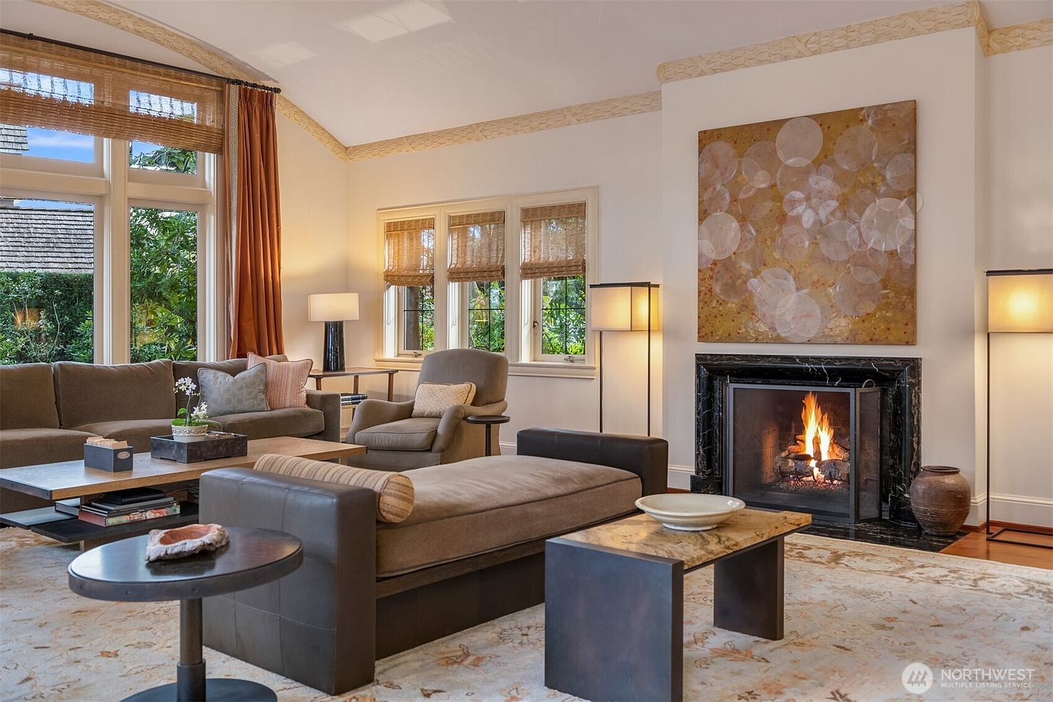 This elegant living room features a vaulted ceiling, a large fireplace with a black marble surround, and a prominent abstract painting. The space is furnished with a comfortable sofa, a chaise lounge, and a patterned area rug, creating a sophisticated and inviting atmosphere. Large windows with woven shades allow for ample natural light, highlighting the warm, neutral color palette and refined decor.