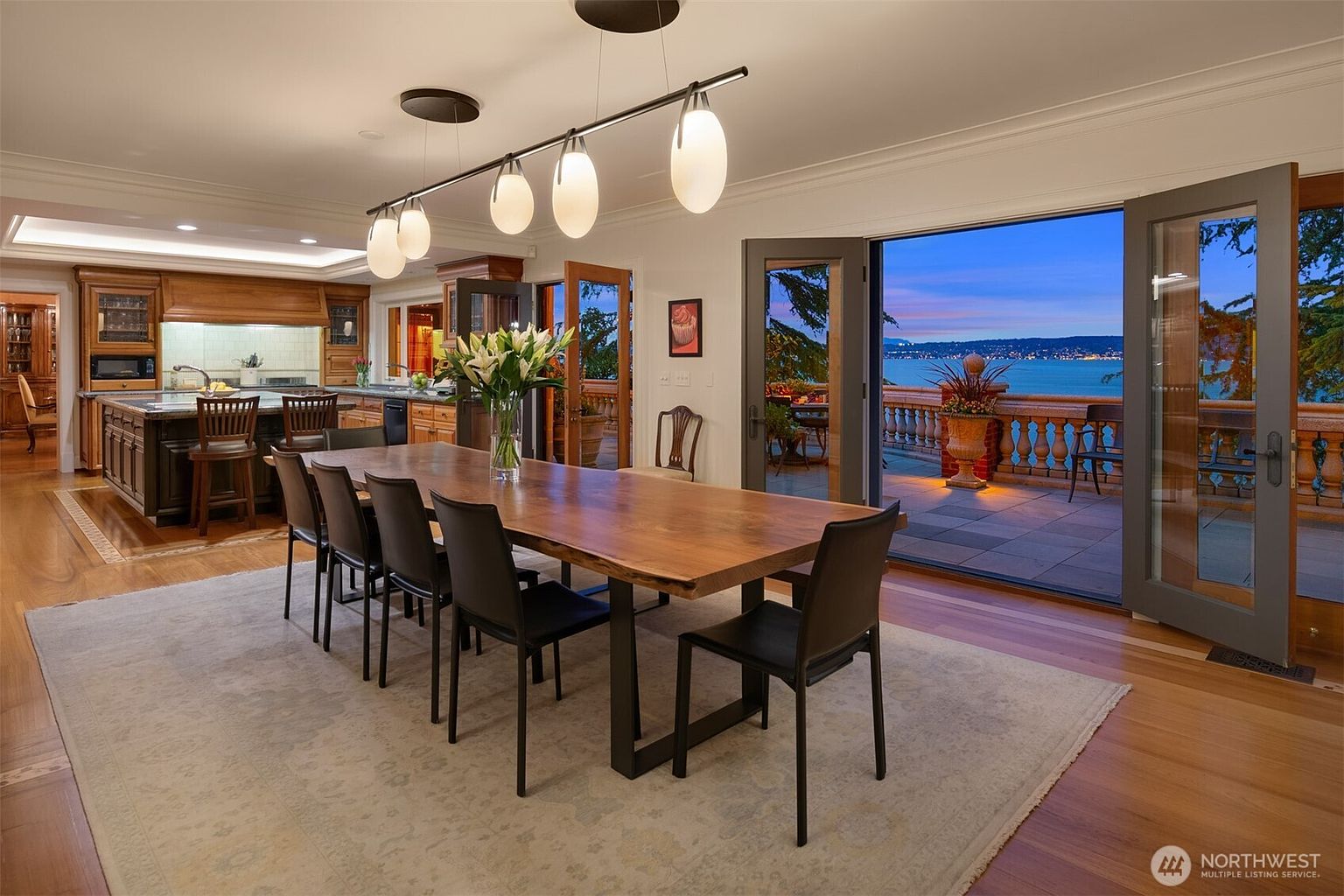 This elegant dining room features a large, natural-edge wood table surrounded by modern black chairs, set atop a neutral area rug. The space flows seamlessly into an open-concept kitchen and opens through wide French doors to a spacious stone patio overlooking a scenic waterfront at dusk. The warm interior lighting contrasts beautifully with the cool, twilight sky visible through the expansive glass doors.