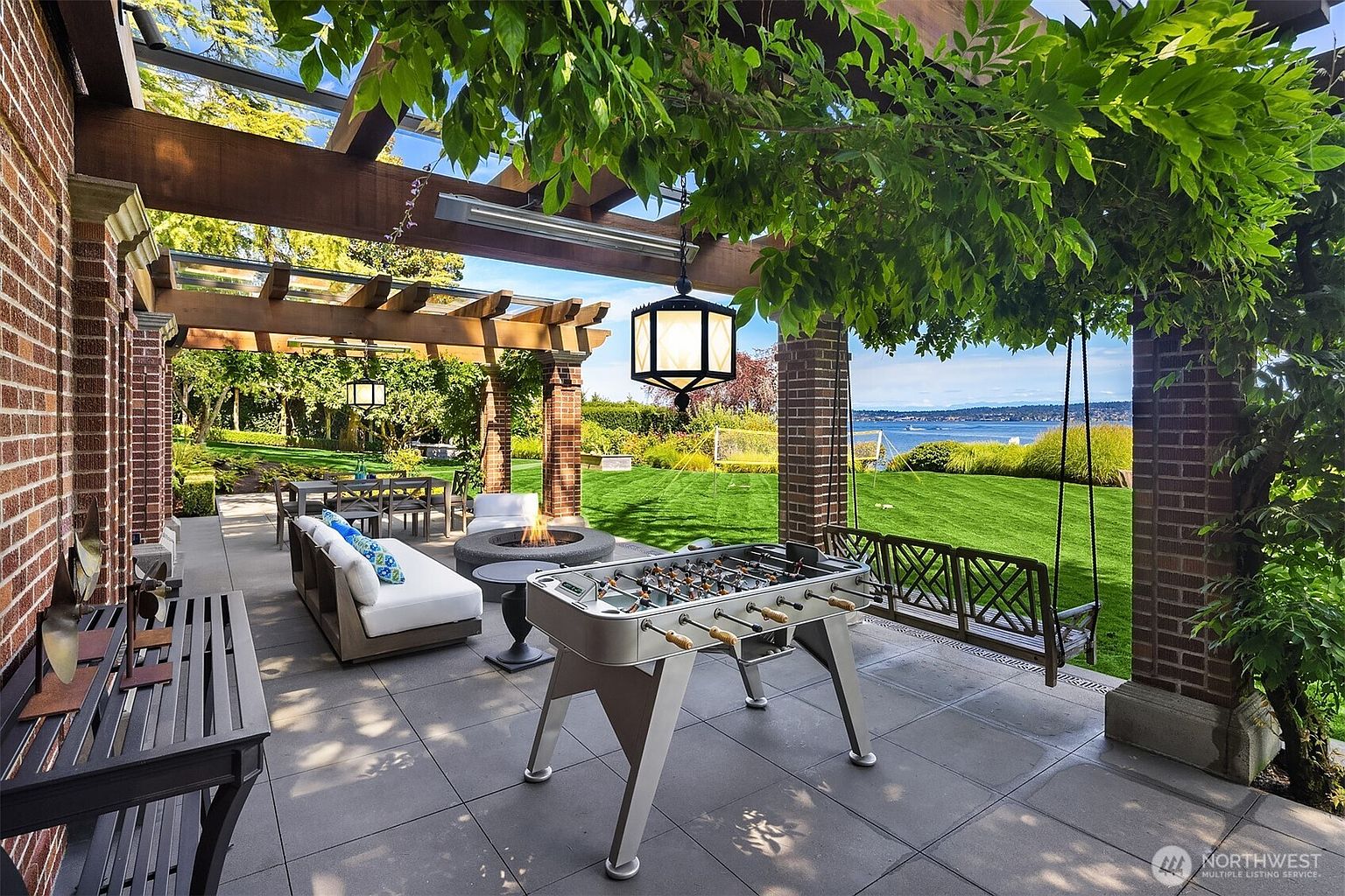This expansive covered patio offers a luxurious outdoor living space, featuring a modern foosball table, a comfortable seating area with a fire pit, and a hanging bench swing. The space is framed by brick pillars and a wooden pergola, providing a seamless transition to a lush green lawn and a stunning waterfront view. The overall atmosphere is sophisticated and perfect for entertaining, blending high-end amenities with a serene natural backdrop.
