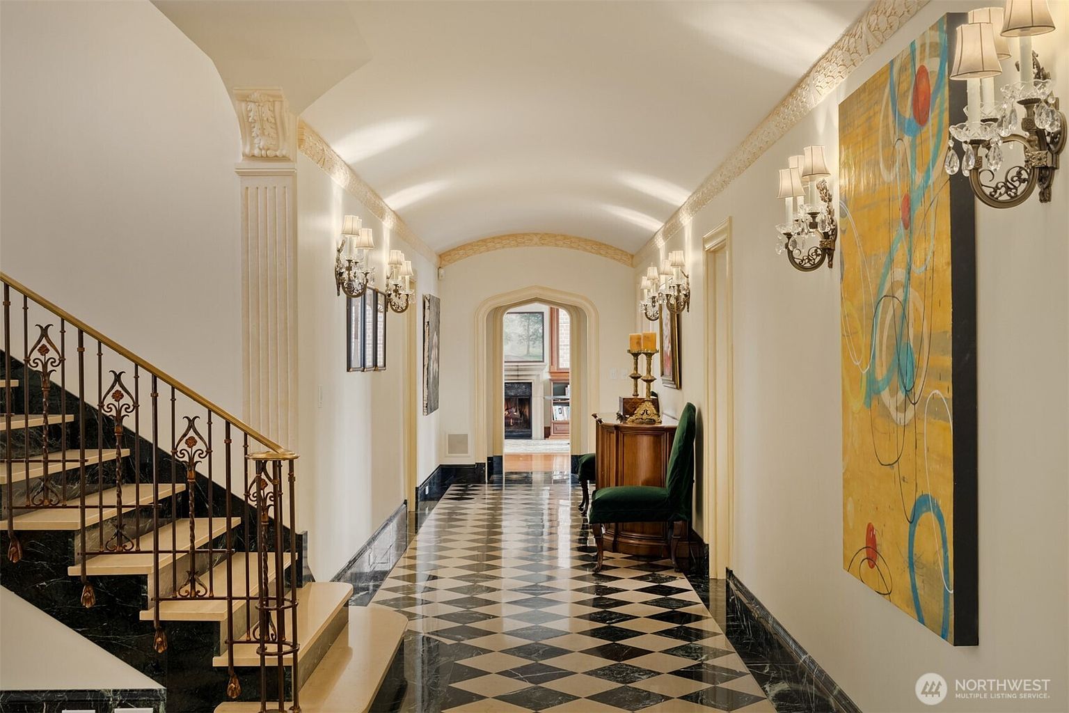 This elegant, grand hallway features a striking black-and-white checkered marble floor and a barrel-vaulted ceiling, creating a sophisticated and luxurious atmosphere. To the left, a sweeping staircase with ornate iron railings leads upward, while the right wall is adorned with a large, vibrant abstract painting and classic wall sconces. The perspective looks down the long corridor toward an arched doorway that reveals a glimpse of a cozy living space with a fireplace, emphasizing the home's spacious and refined architectural design.
