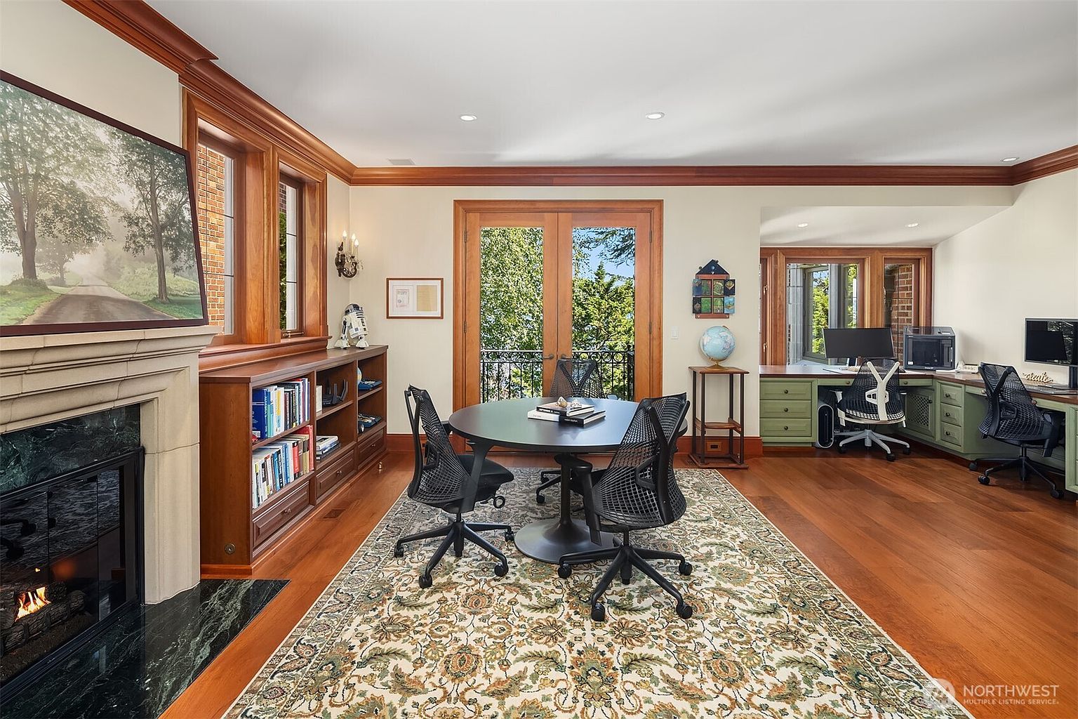 This sophisticated home office features rich wood trim, built-in bookshelves, and a cozy fireplace, creating a warm and professional atmosphere. A central round table with ergonomic chairs serves as a collaborative workspace, while a dedicated desk area with green cabinetry provides a functional workstation. The room is bathed in natural light from the large windows and French doors, which open onto a balcony, offering a seamless blend of indoor and outdoor living.