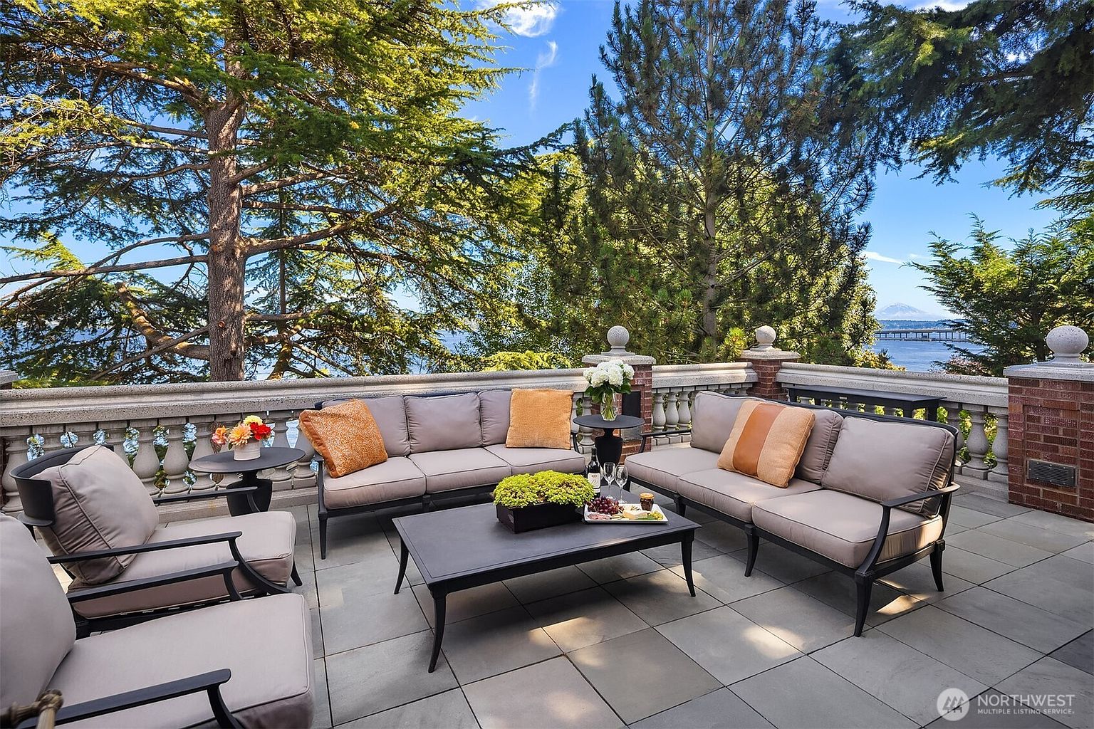 This elegant outdoor terrace features a sophisticated seating arrangement with plush, neutral-toned sofas and armchairs centered around a sleek, dark metal coffee table. The space is framed by a classic stone balustrade and brick pillars, offering a stunning, elevated view of the water, distant mountains, and lush surrounding trees. The overall atmosphere is one of refined relaxation, perfect for entertaining or enjoying a peaceful afternoon with a scenic backdrop.