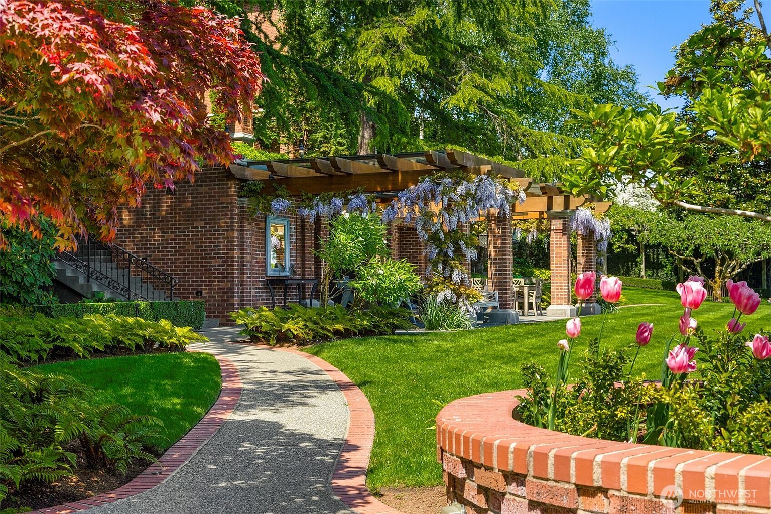 This picturesque garden scene features a winding stone pathway leading toward a charming brick-clad home with a wooden pergola covered in blooming wisteria. The lush, manicured lawn is framed by vibrant pink tulips in a curved brick planter and a variety of mature trees and ferns, creating a serene and elegant outdoor living space. The perspective is a ground-level shot that emphasizes the depth and inviting atmosphere of the backyard landscape.