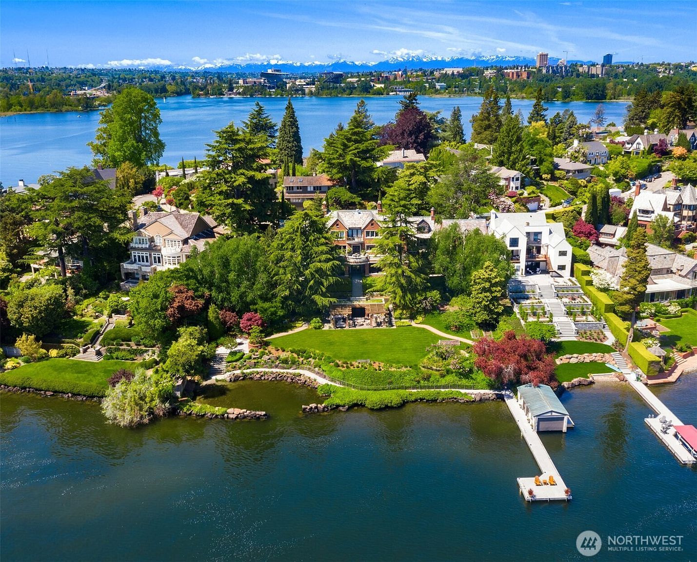 This stunning aerial view captures a luxurious waterfront estate nestled along a serene lake, featuring expansive manicured lawns, tiered gardens, and a private dock. The property showcases a grand multi-story residence with classic architectural details, surrounded by lush mature trees and vibrant landscaping. The perspective provides a comprehensive look at the home's prime location, highlighting its seamless integration with the natural beauty of the shoreline and the distant mountain backdrop.