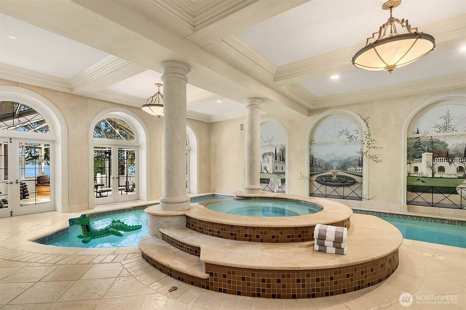 This luxurious indoor pool and spa area features a grand, circular raised hot tub as the centerpiece, flanked by elegant columns and surrounded by a tiled pool. The room is adorned with artistic wall murals, arched glass doors leading to an outdoor view, and sophisticated chandelier lighting, creating a serene and opulent atmosphere. The perspective is a wide, eye-level shot that captures the symmetry and high-end finishes of this unique recreational space.