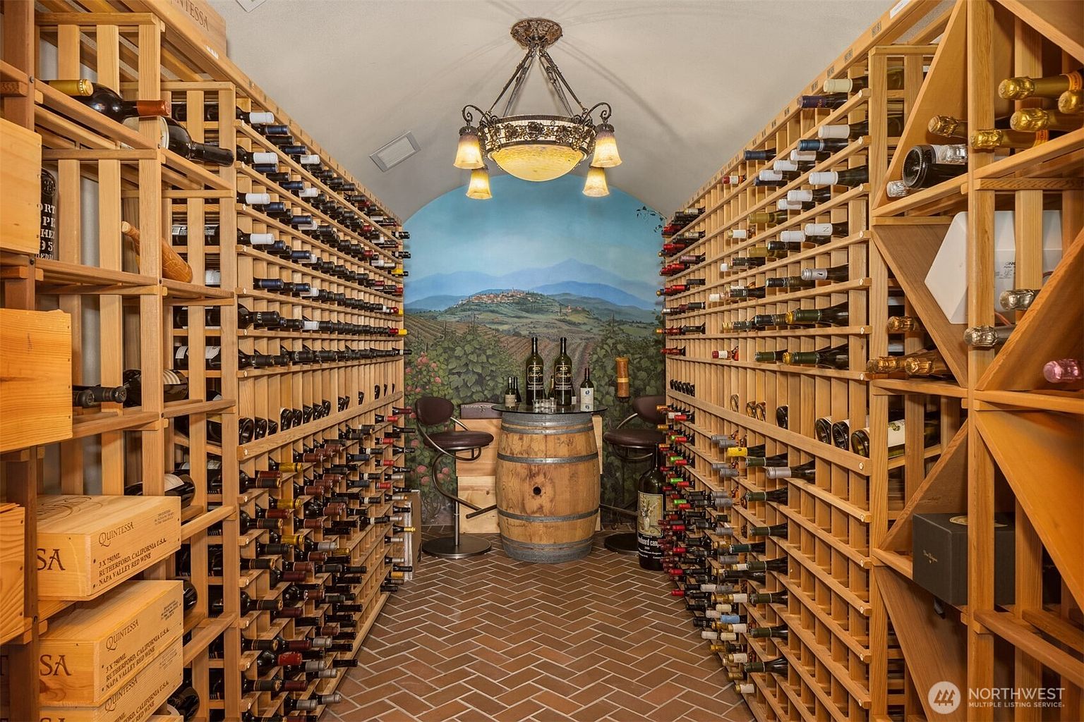 This sophisticated wine cellar features floor-to-ceiling wooden racking systems filled with an extensive collection of wine bottles, creating an immersive and luxurious atmosphere. A central barrel-top table serves as a focal point, complemented by a scenic mural of a vineyard landscape and an ornate chandelier overhead. The space is finished with a classic herringbone-patterned brick floor, evoking the charm of a traditional European winery.