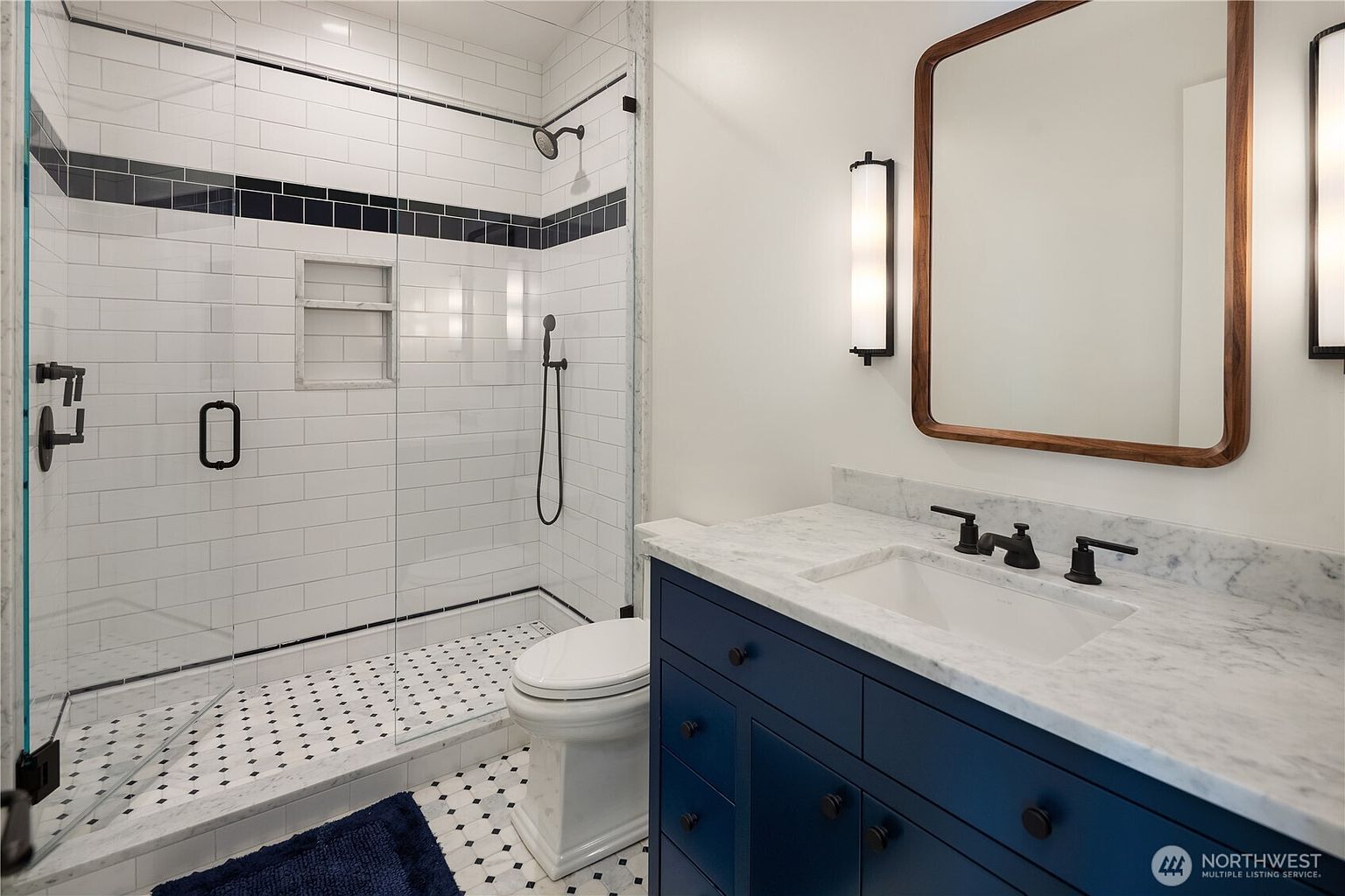 This modern bathroom features a striking navy blue vanity topped with a light-colored stone counter and a rectangular undermount sink. To the left, a glass-enclosed walk-in shower showcases white subway tile walls with a dark accent stripe, a built-in niche, and a handheld showerhead. The space is completed by a rounded rectangular mirror, wall-mounted lighting, and a classic black-and-white patterned tile floor.