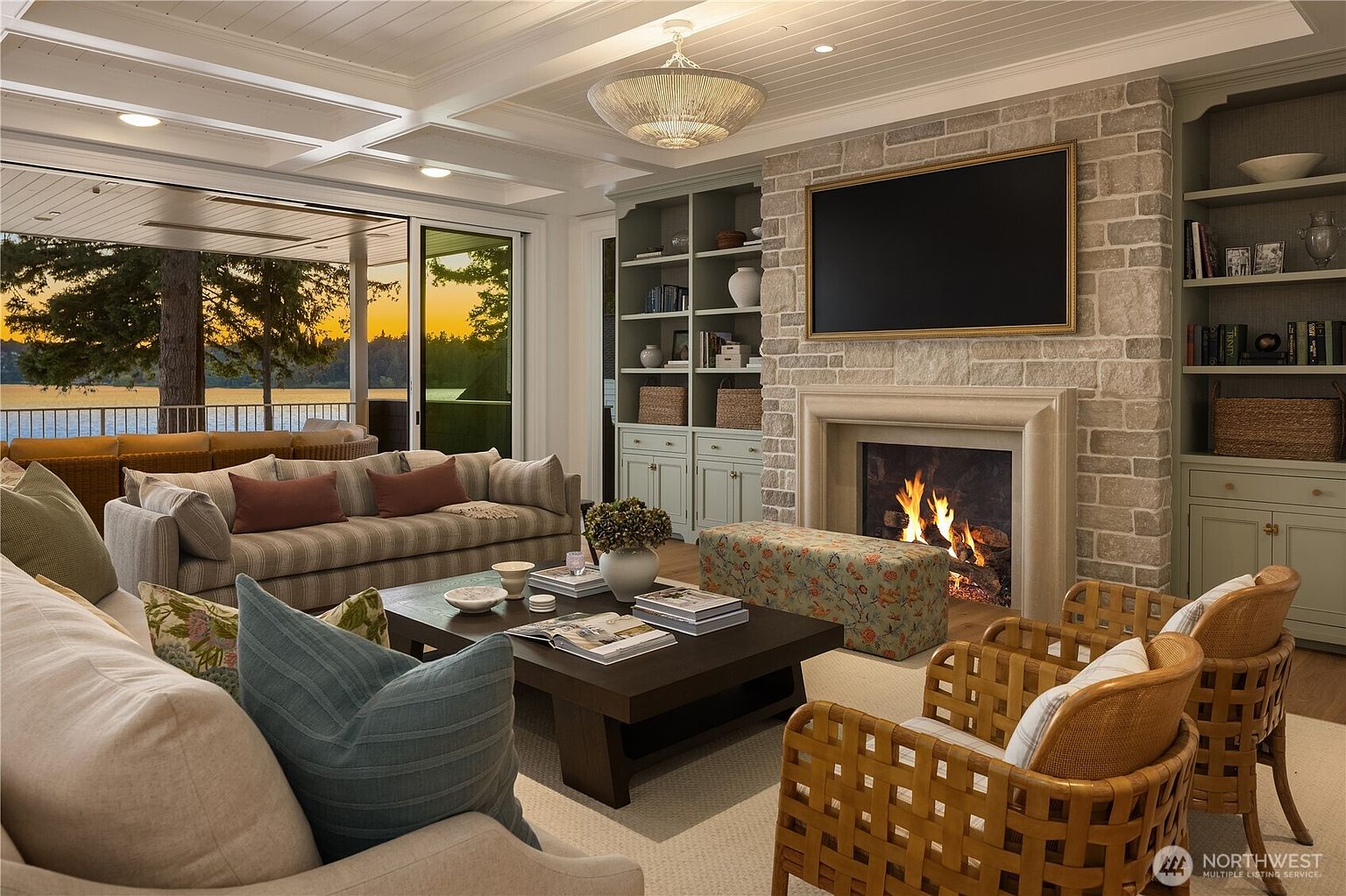 This elegant living room features a stone-clad chimney breast surrounding a cozy fireplace, flanked by built-in cabinetry in a soft sage tone. A large, plush striped sofa and woven armchairs create a comfortable seating area centered around a dark wood coffee table, while expansive sliding glass doors open to a covered deck with a scenic sunset view over the water. The space is illuminated by a decorative chandelier and recessed lighting, offering a sophisticated and inviting atmosphere.