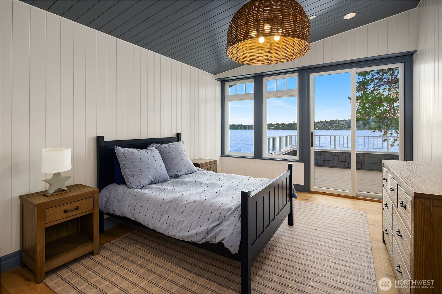 This serene bedroom features a dark-framed bed with patterned bedding, flanked by light wood nightstands and a matching dresser. The room is accented by a dark-painted wood-paneled ceiling and a large woven pendant light, while a sliding glass door provides direct access to a balcony with expansive water views. The space is grounded by a neutral striped area rug, creating a calm and inviting atmosphere.