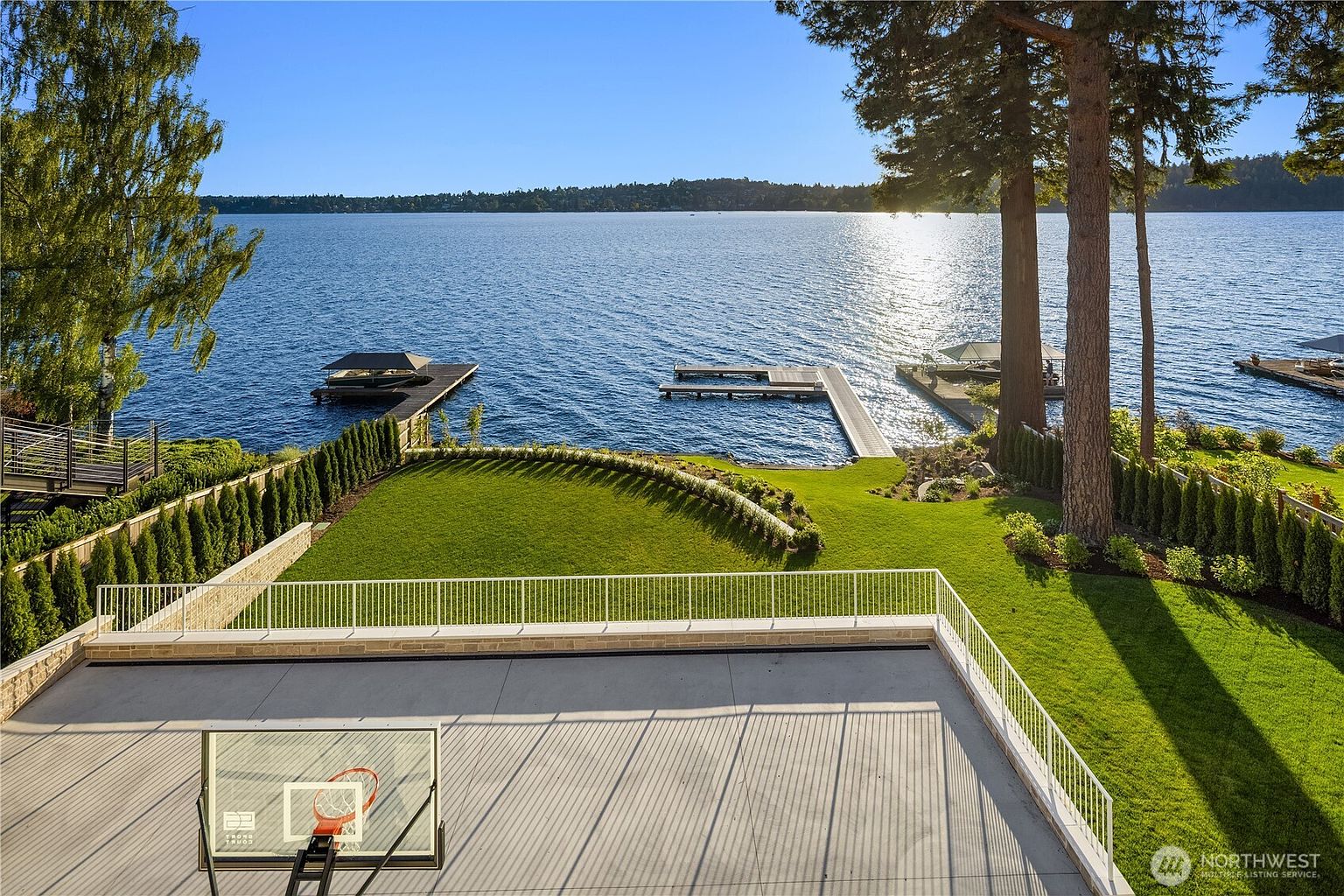 This elevated perspective captures a sprawling backyard overlooking a serene lake, featuring a paved sports court with a basketball hoop in the foreground. A manicured lawn slopes gently toward the water, bordered by a row of tall, slender evergreen trees and a white railing. The scene includes private docks extending into the lake, framed by mature trees and a clear blue sky, creating a peaceful and luxurious waterfront atmosphere.