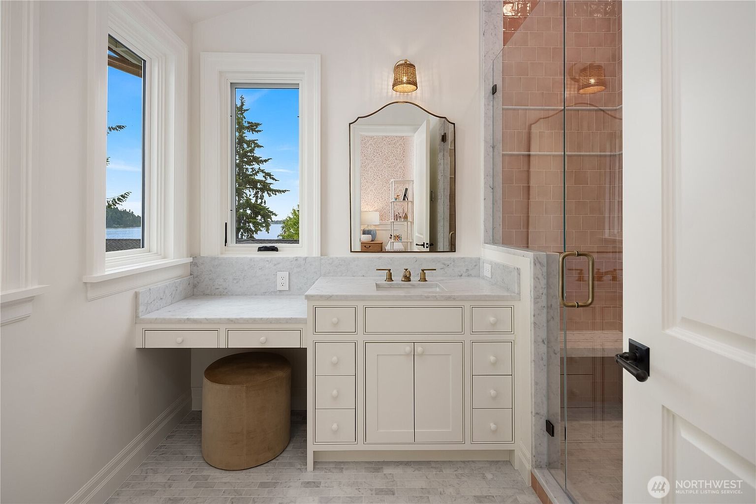 This bright and elegant bathroom features a white vanity with a stone countertop, a matching makeup desk area, and a large arched mirror. To the right, a glass-enclosed shower is lined with warm-toned rectangular tiles, while two windows provide natural light and a view of the water. The space is finished with light-colored stone flooring and a neutral color palette, creating a clean and sophisticated atmosphere.