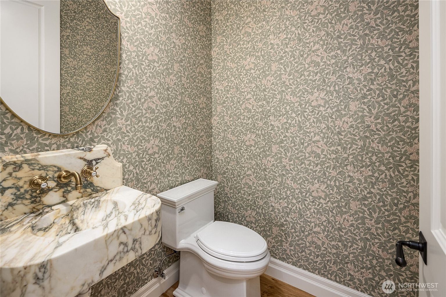 This powder room features a distinctive stone vanity with a prominent, swirling pattern and gold-toned fixtures. The walls are adorned with a detailed, nature-inspired botanical wallpaper in muted green and pink tones, complemented by a round mirror with a thin gold frame. A white toilet is positioned against the patterned wall, creating a cohesive and elegant aesthetic for a guest space.