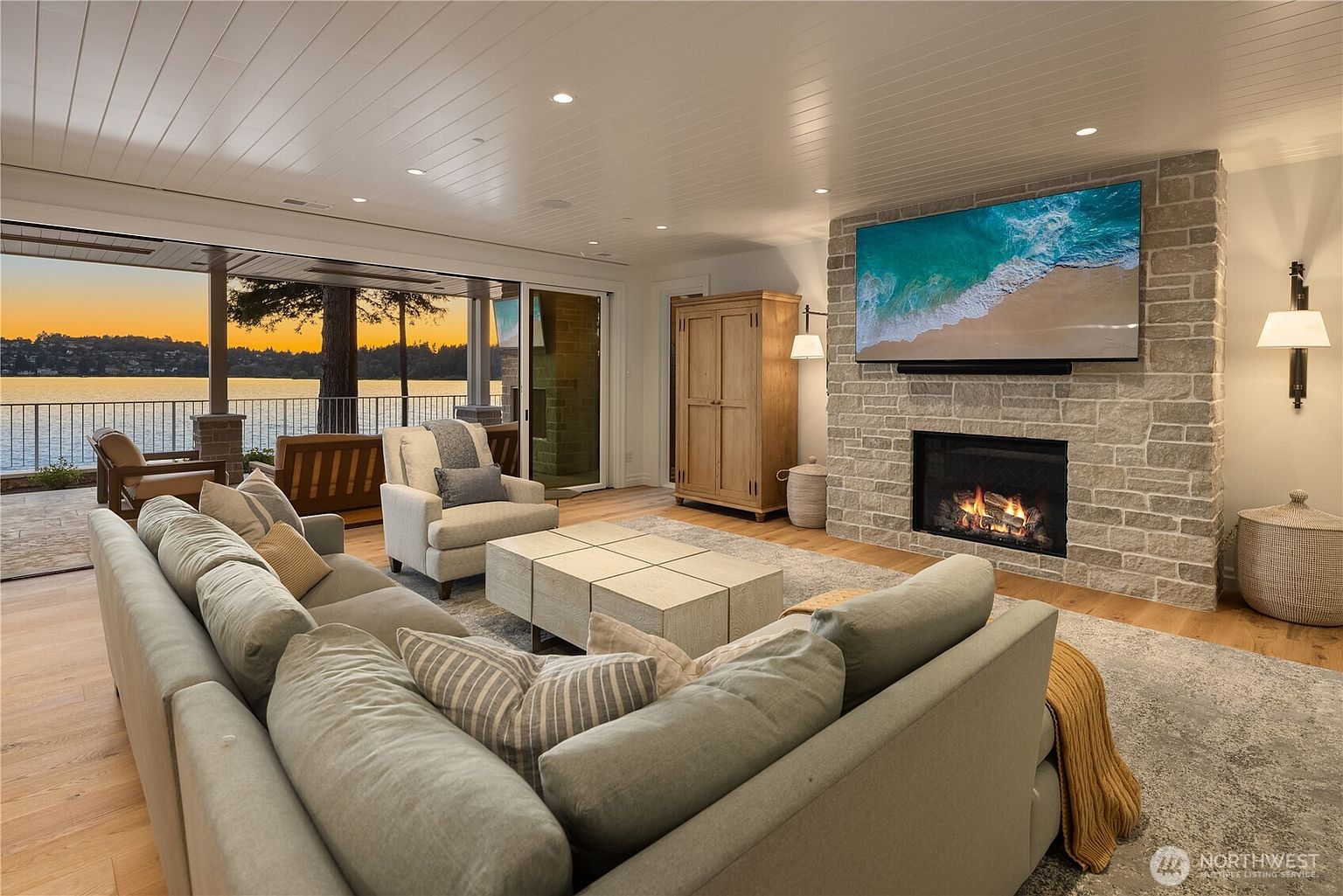 This spacious living room features a large stone-clad chimney breast housing a fireplace and a mounted television, serving as the room's focal point. The space is furnished with a plush sectional sofa, a light-toned coffee table, and an armchair, all arranged on a textured area rug. Large sliding glass doors open to a covered deck, providing an expansive view of the lake at sunset and creating a seamless indoor-outdoor living experience.