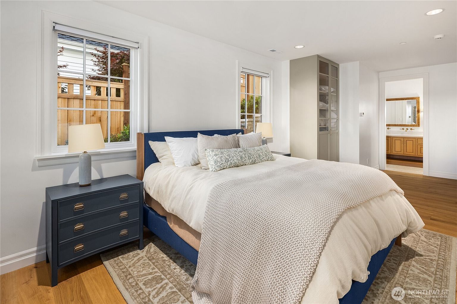 This bright and airy bedroom features a central bed with a navy blue upholstered frame, dressed in neutral linens and a textured knit throw. A matching navy blue nightstand sits to the left, topped with a modern table lamp, while a tall, light-toned storage cabinet stands against the back wall. The room is illuminated by natural light from two windows and leads into an adjacent bathroom, creating a cohesive and inviting living space.