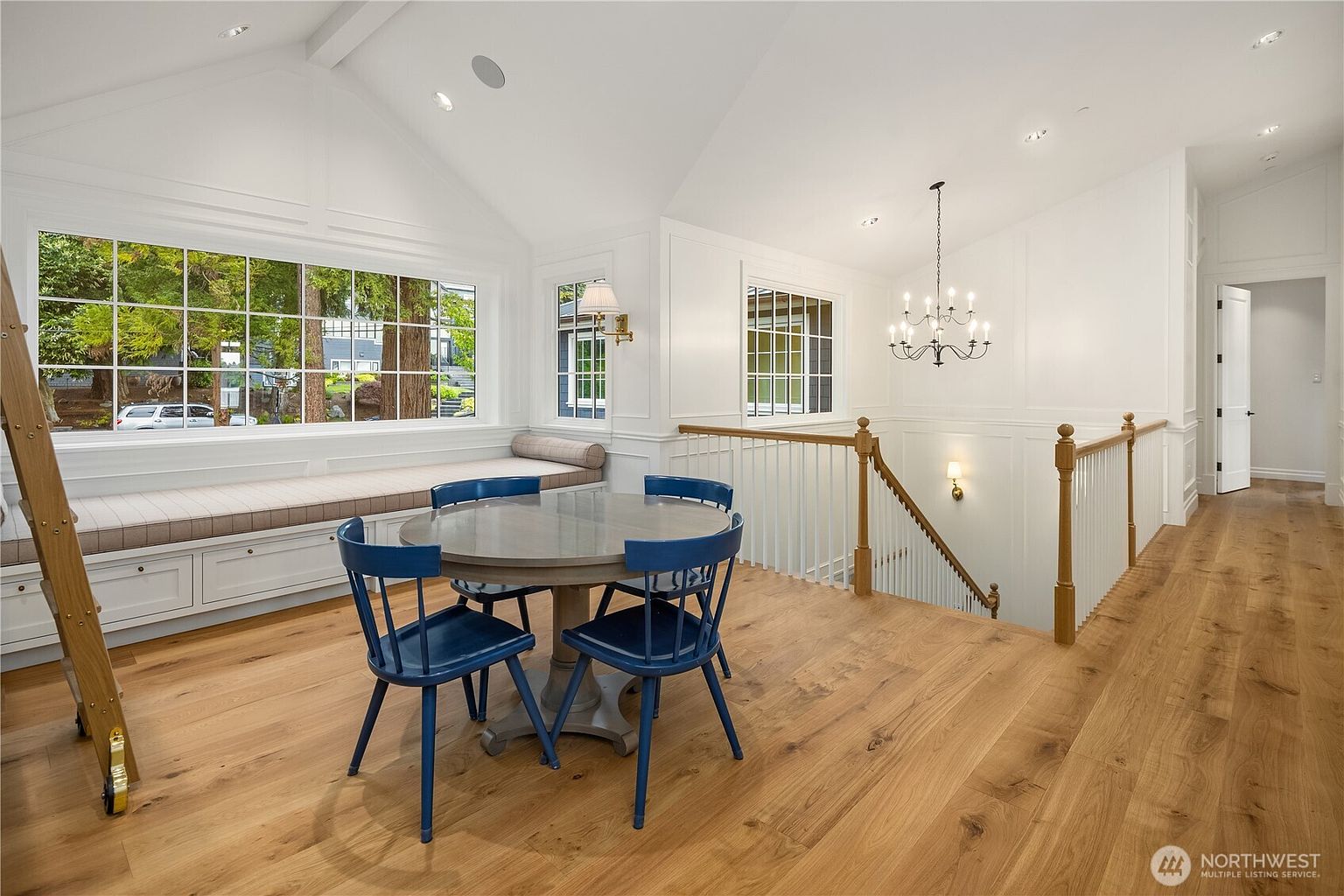 This bright, open landing area features light wood flooring and a vaulted ceiling, creating an airy and spacious atmosphere. A built-in window seat with a cushioned bench sits beneath a large window, while a round table with four blue chairs provides a cozy seating nook. The space overlooks a staircase with a wooden railing and is illuminated by a decorative chandelier and recessed lighting.