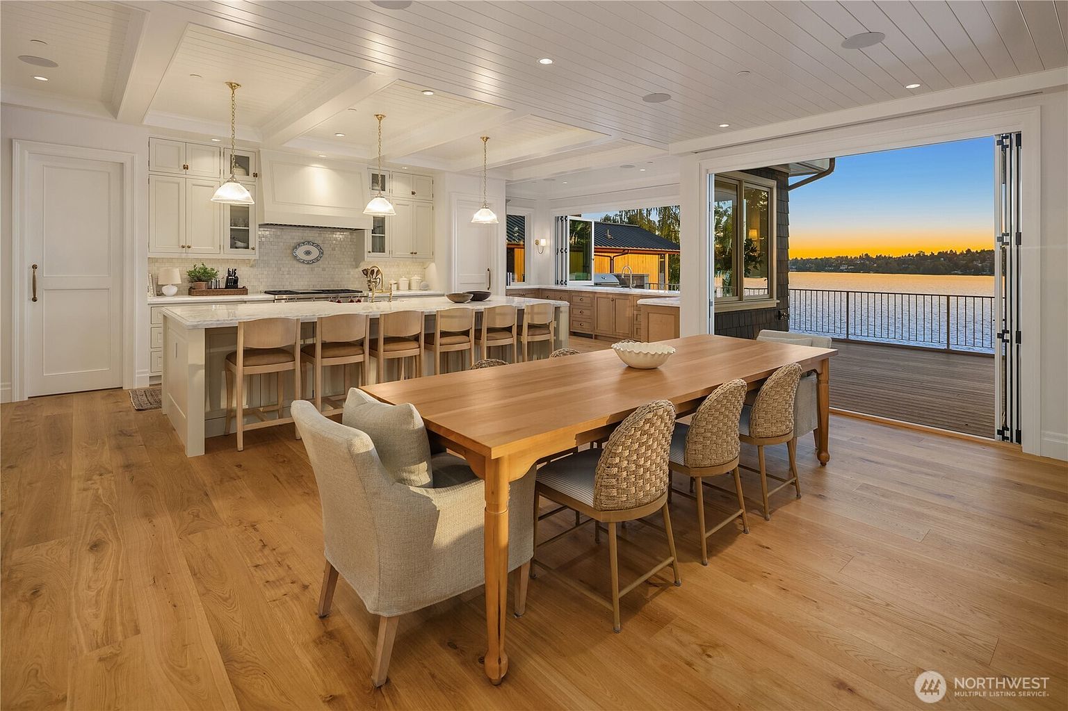 This bright, open-concept kitchen and dining area features a large central island with seating, light wood flooring, and white cabinetry. A long wooden dining table sits in the foreground, while a set of folding glass doors opens completely to a deck overlooking a serene lake at sunset. The space is illuminated by pendant lighting and recessed ceiling lights, creating an airy and inviting atmosphere.