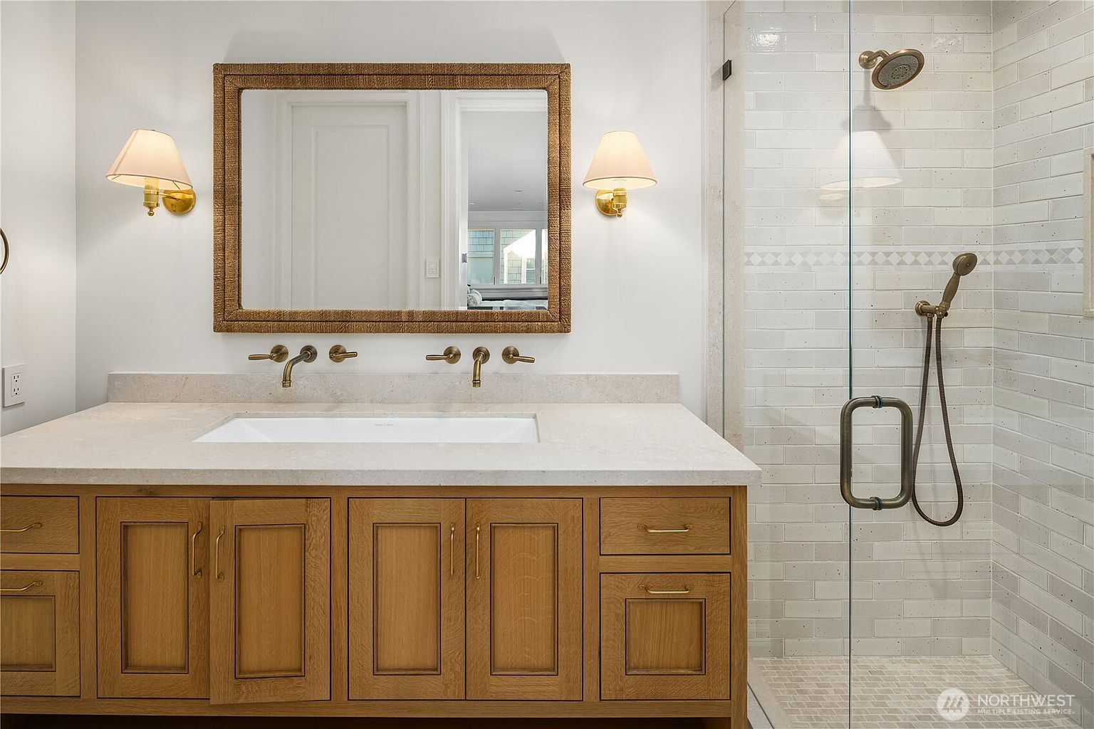 This bright and clean bathroom features a light wood vanity with a stone countertop and a rectangular undermount sink. Above the vanity, a textured-frame mirror is flanked by two wall-mounted sconces, while a glass-enclosed shower with light-colored subway tile and a handheld showerhead sits to the right. The space is well-lit and maintains a neutral, modern aesthetic.