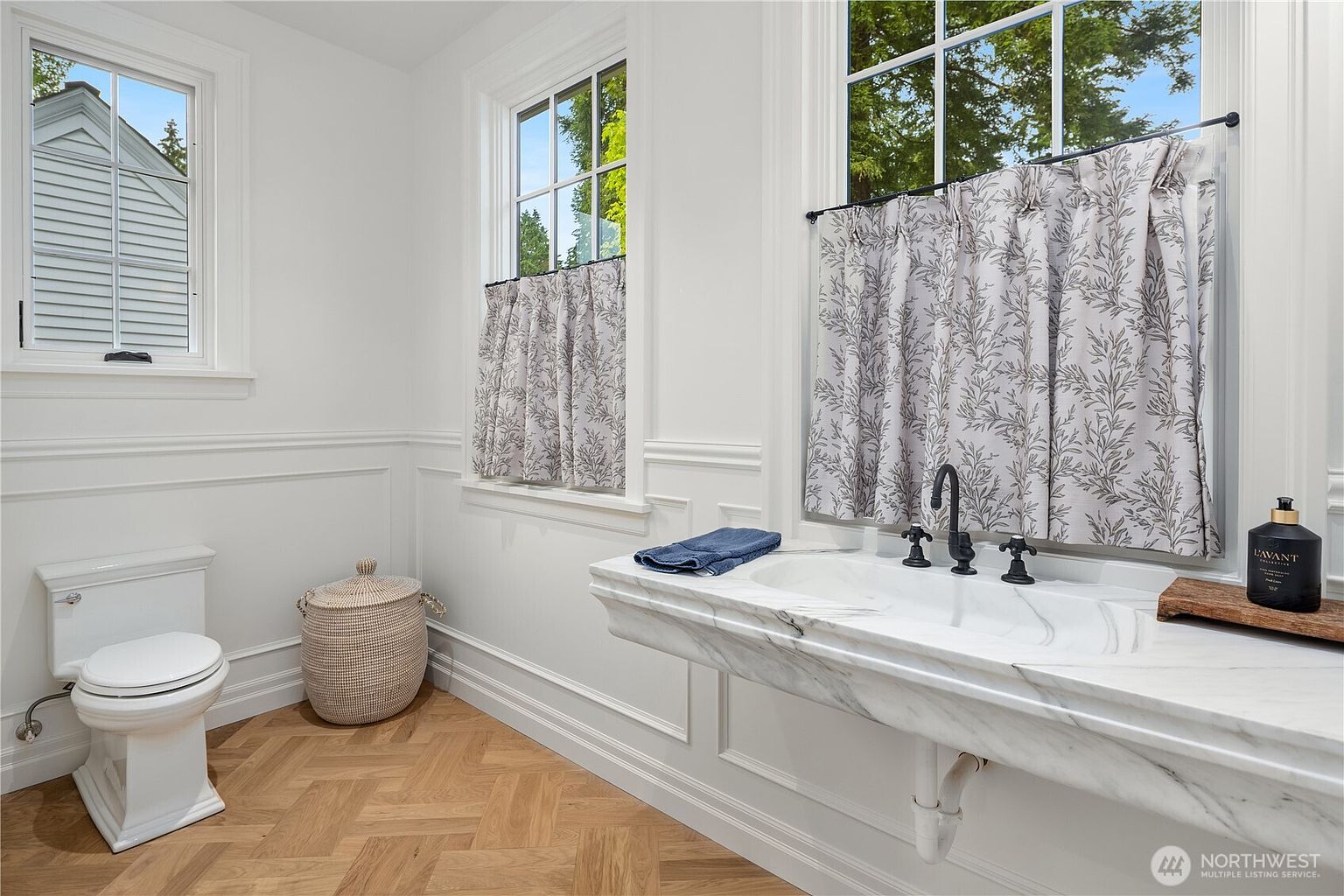 This bright powder room features a distinctive wall-mounted stone sink with a prominent veined pattern and dark hardware. The space is finished with crisp white wainscoting, patterned fabric curtains on the windows, and light-toned wood flooring laid in a herringbone pattern. A white toilet and a woven storage basket complete the clean, elegant aesthetic of this small bathroom.