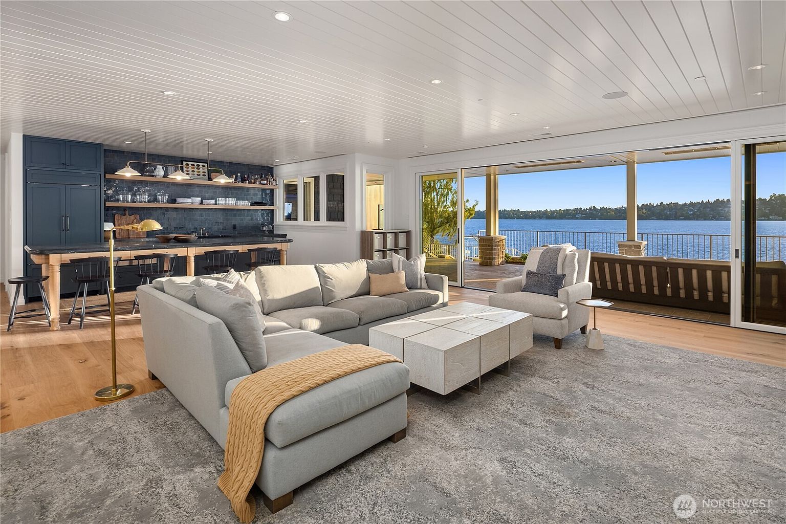 This open-concept living space features a large sectional sofa and a central coffee table, seamlessly transitioning into a kitchen area with dark cabinetry and a breakfast bar. The room is defined by its expansive floor-to-ceiling sliding glass doors that open to a waterfront deck, offering panoramic views of the lake. The interior aesthetic is bright and airy, characterized by light wood flooring and a clean, modern design.