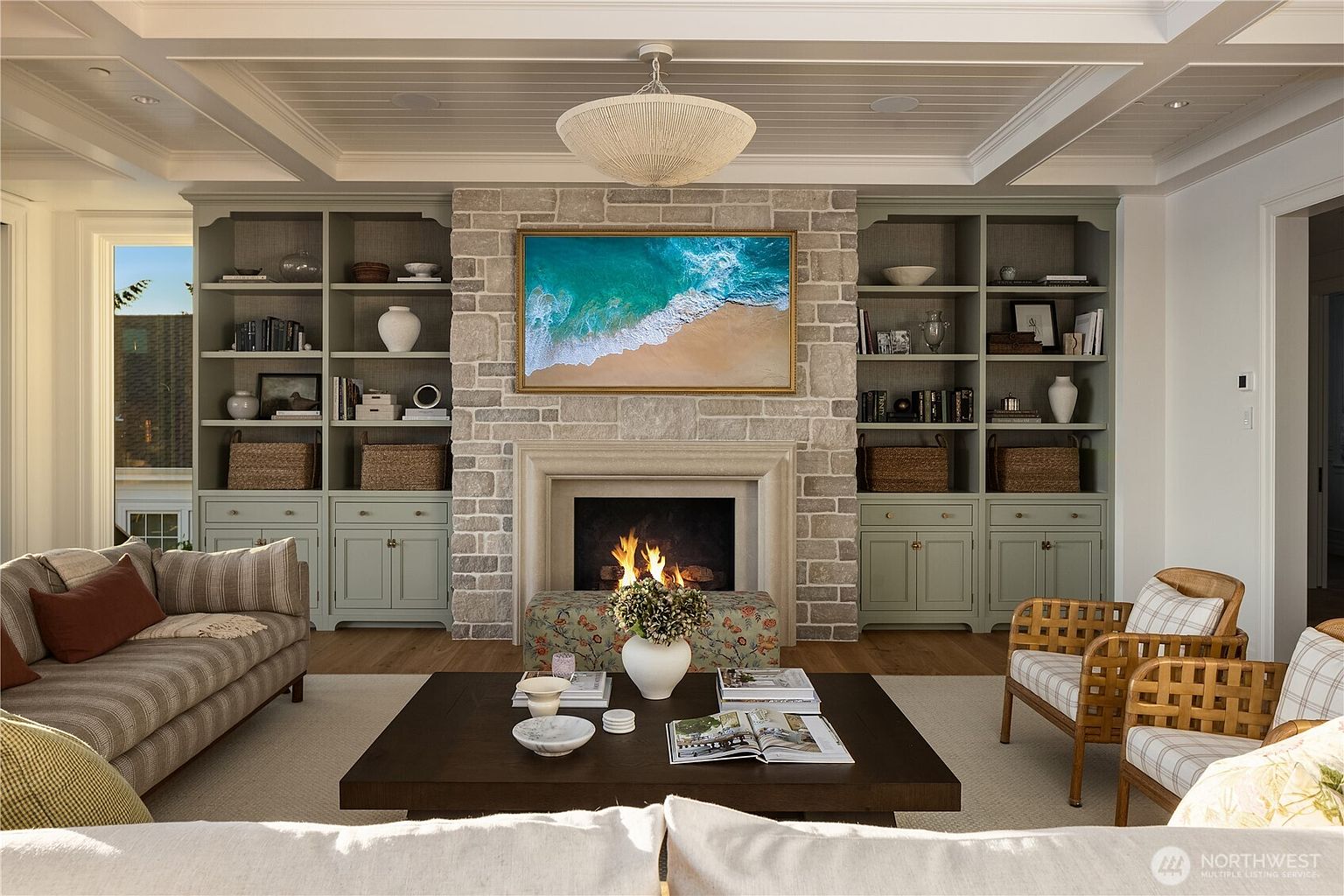 This elegant living room features a central stone-clad chimney breast housing a fireplace and a television displaying a coastal scene. Flanking the fireplace are custom built-in cabinets in a soft sage green, providing ample display and storage space. The room is anchored by a dark wood coffee table and a neutral area rug, creating a sophisticated and inviting atmosphere.
