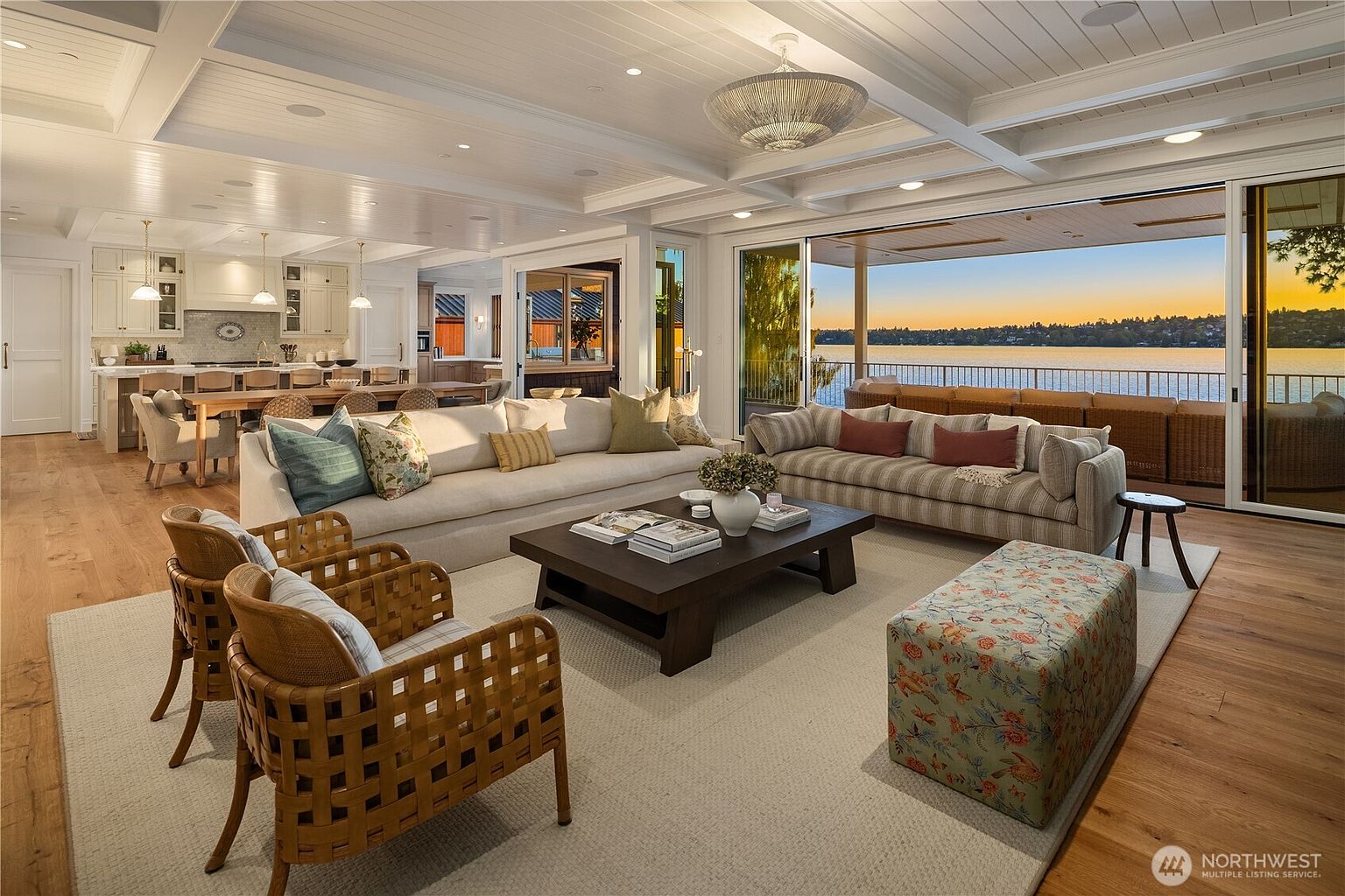 This expansive, open-concept living area features a sophisticated coastal aesthetic with light wood flooring and a coffered ceiling. The space is anchored by a large, neutral-toned sofa and a dark wood coffee table, while floor-to-ceiling sliding glass doors open to a covered deck overlooking a serene lake at sunset. A kitchen with a long island and bar seating is visible in the background, creating a seamless flow for entertaining.