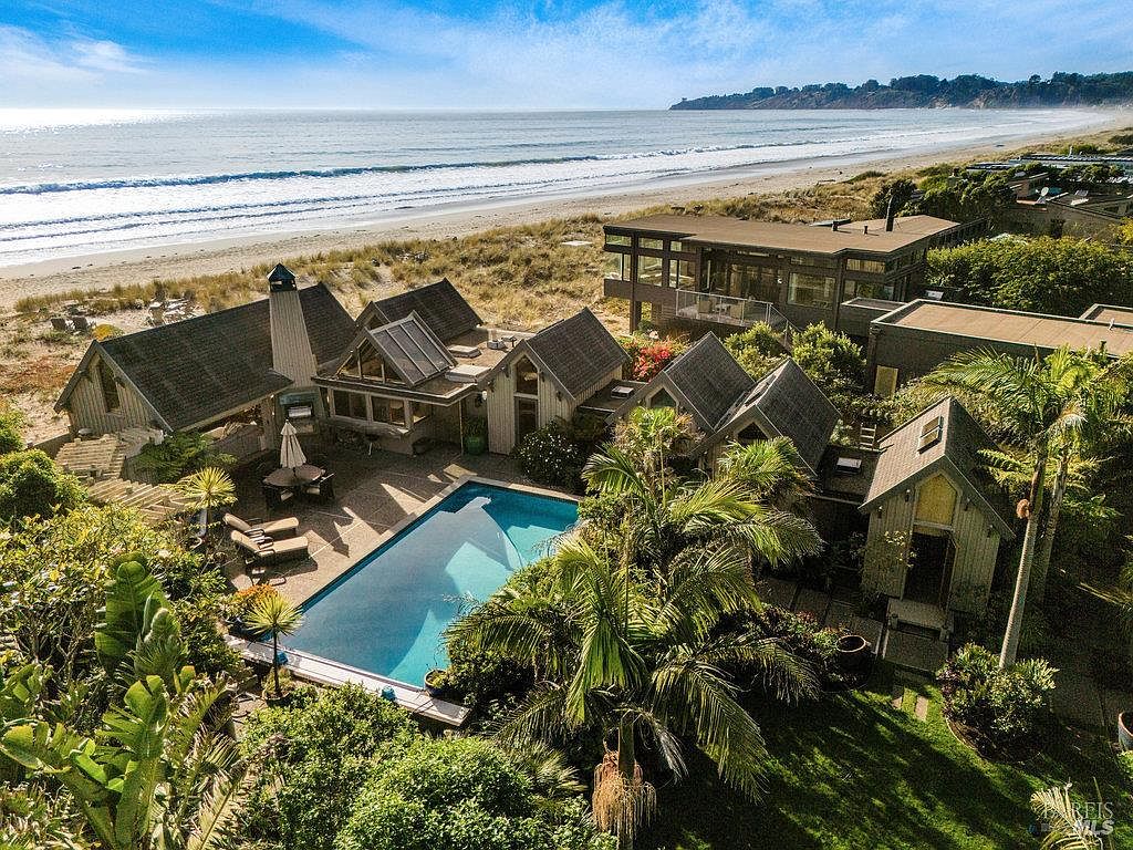 This high-angle aerial view captures a coastal property featuring a collection of interconnected, cottage-style structures with shingled roofs. A rectangular swimming pool serves as the centerpiece of the courtyard, surrounded by lush tropical landscaping and a paved patio area. The property is situated directly adjacent to a sandy beach and the ocean, offering a serene and private retreat with a distinct architectural character.