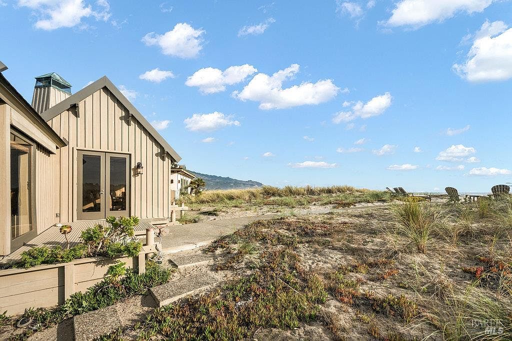 This rear view of a coastal home showcases vertical wood siding and a set of glass-paned double doors leading onto a wooden deck. A concrete path winds through the sandy, dune-like landscape, which is dotted with native grasses and low-lying vegetation. The scene captures a serene beachside atmosphere with a clear blue sky and distant hills in the background.