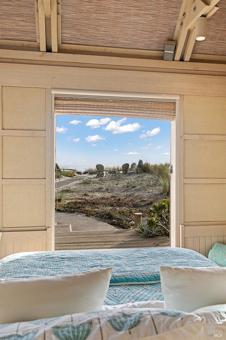 This bedroom features a serene, coastal aesthetic with light-toned wood paneling on the walls and a vaulted ceiling. A large, open doorway frames a direct view of a sandy beach, dunes, and outdoor seating, seamlessly blending the indoor space with the natural landscape. The foreground includes a neatly made bed with a blue and white patterned quilt, creating a tranquil and inviting atmosphere.