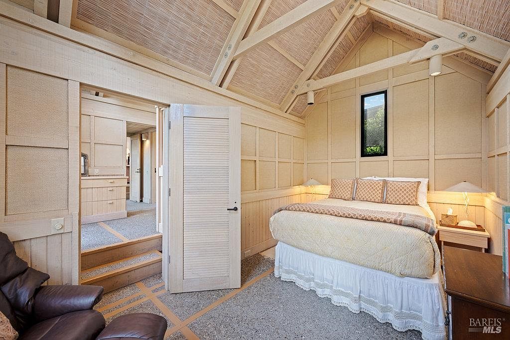 This cozy bedroom features a vaulted ceiling with exposed beams and light-toned wood paneling throughout the walls. A queen-sized bed with a patterned quilt is positioned against the back wall, flanked by two small bedside lamps, while a leather armchair sits in the foreground. A doorway leads to an adjacent hallway, highlighting the home's rustic, cabin-like aesthetic and warm, inviting atmosphere.