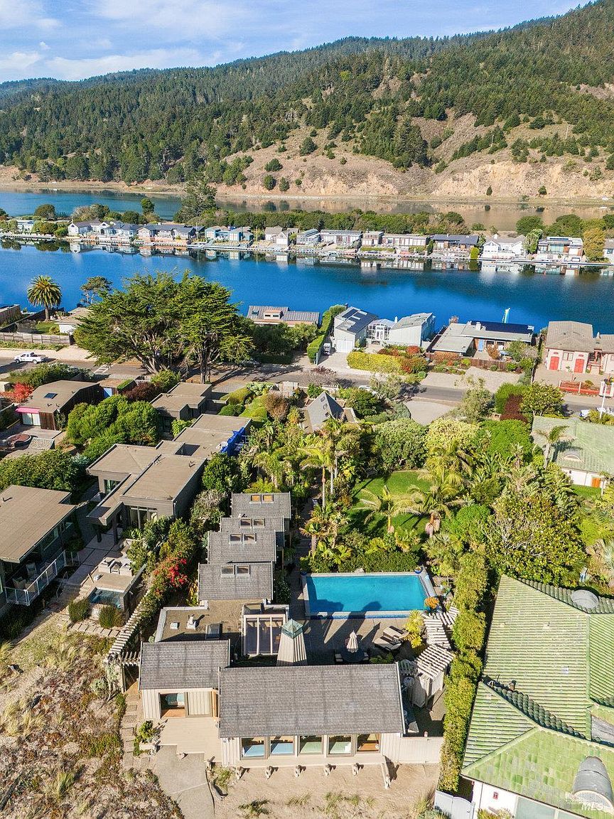 This high-angle aerial view captures a coastal residential property nestled between a serene blue waterway and a lush, tree-covered hillside. The home features a multi-sectioned roofline, a private rectangular swimming pool set within a landscaped yard, and a series of interconnected structures. The surrounding neighborhood is densely populated with similar waterfront homes, creating a tranquil and scenic community atmosphere.