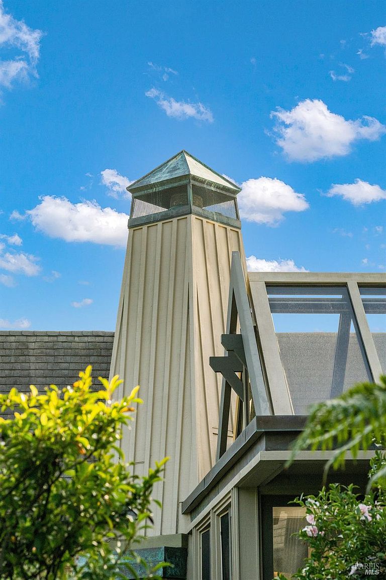 This image captures a unique, tapered chimney structure with vertical siding, topped by a copper-capped vent with protective mesh screening. To the right, a portion of a glass-paneled roof structure is visible, framed by dark structural beams. The composition is set against a bright blue sky with scattered clouds, highlighting the architectural geometry of the home's exterior.
