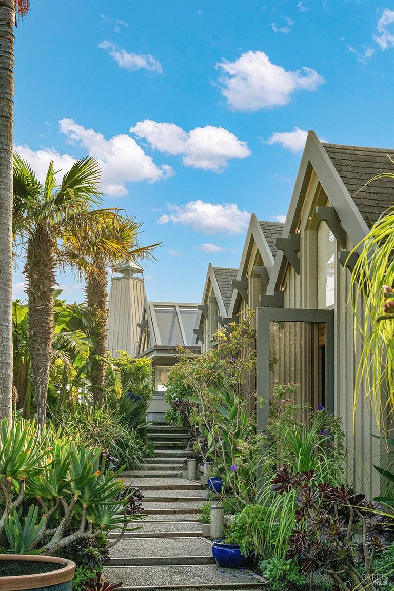 This exterior view captures a lush, landscaped garden path leading toward a series of modern, gabled structures with vertical siding. The walkway is composed of concrete steps and gravel, flanked by vibrant tropical plants, palm trees, and succulents that create a serene, resort-like atmosphere. The perspective is a low-angle shot looking up the path, emphasizing the architectural rhythm of the rooflines against a bright blue sky with scattered clouds.