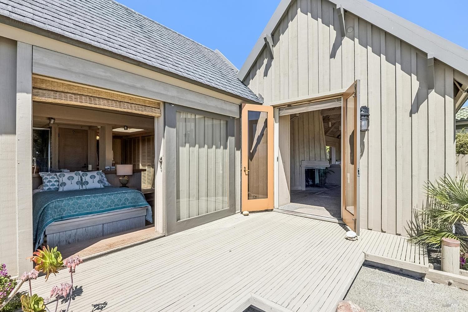 This image captures a bright, coastal-style outdoor deck that seamlessly connects to the interior living spaces through wide-opening glass doors. The deck features light-toned wood planking and leads directly into a bedroom on the left and a living area with a visible fireplace on the right. The architectural style is characterized by vertical wood siding and a clean, airy aesthetic that emphasizes indoor-outdoor living.