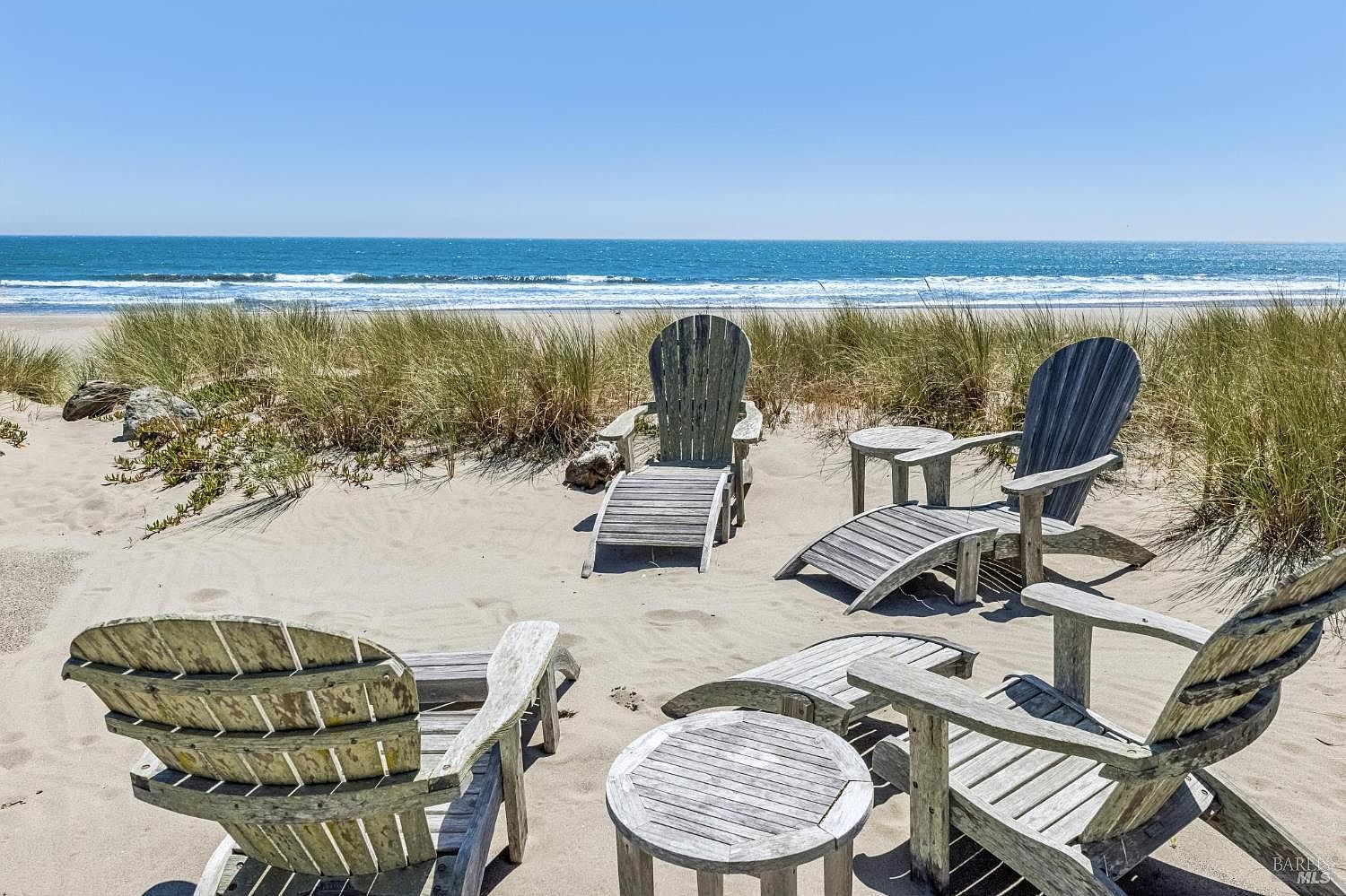 This serene outdoor seating area is situated directly on a sandy beach, featuring several weathered wooden Adirondack chairs and matching side tables. The arrangement is nestled among natural dune grasses, providing a peaceful vantage point overlooking the vast blue ocean and horizon. The scene captures a relaxed, coastal lifestyle with an unobstructed view of the shoreline.