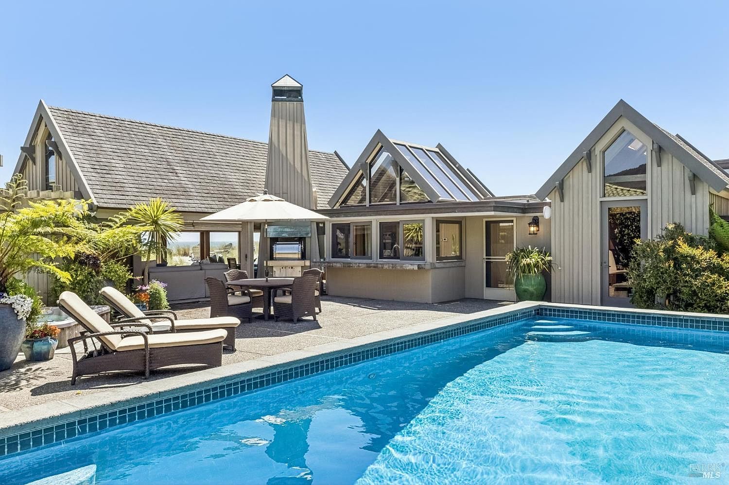 This outdoor living space features a rectangular swimming pool with blue tile trim, set against a multi-gabled home with light-colored siding. A paved patio area includes a dining set under a white umbrella, a built-in outdoor grill, and lounge chairs, creating a relaxing environment. The architectural design incorporates large windows and a prominent chimney, offering a bright and inviting atmosphere for outdoor entertaining.