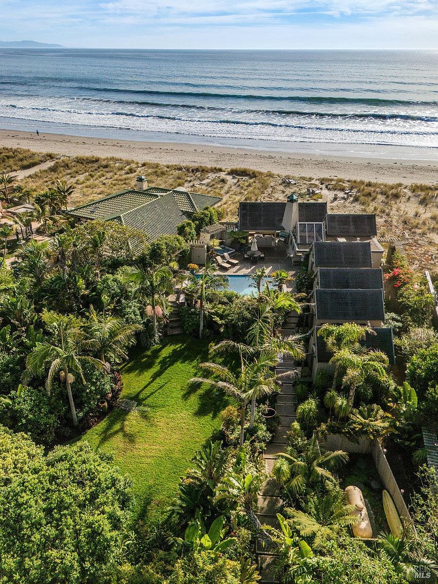 This high-angle aerial view captures a luxurious beachfront property nestled among lush tropical landscaping and mature palm trees. The estate features a private swimming pool, a sprawling green lawn, and a series of connected structures with dark shingled roofs that lead directly to the sandy dunes and the ocean. The perspective emphasizes the seamless integration of the home into its coastal environment, highlighting the expansive outdoor living spaces and direct beach access.