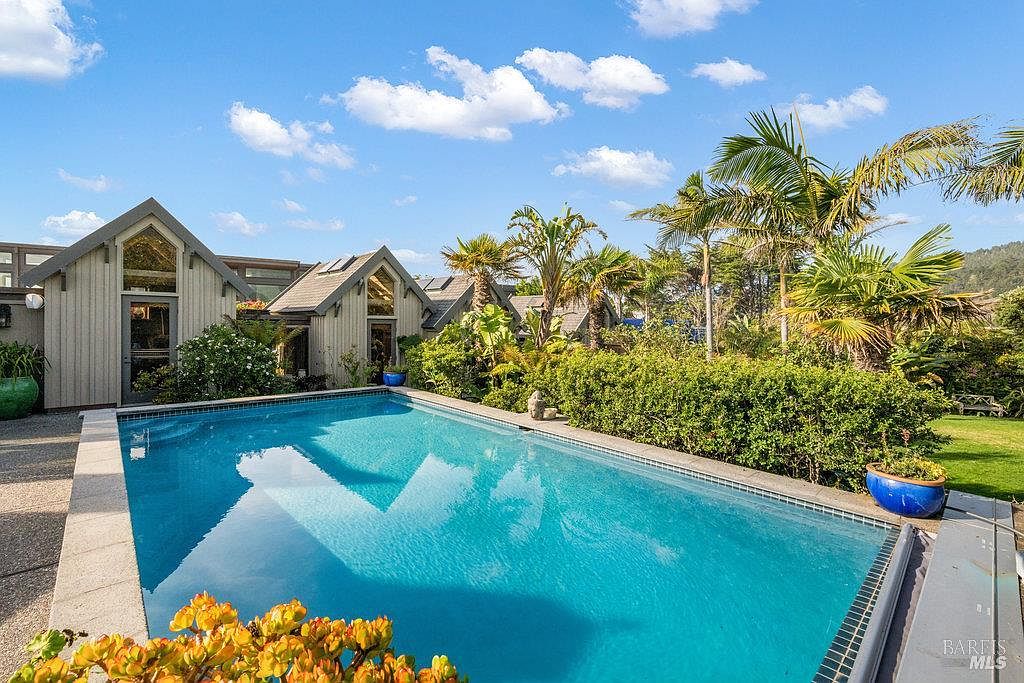 This image captures a serene backyard oasis featuring a rectangular swimming pool with clear blue water, set against a backdrop of lush greenery and palm trees. The home's exterior showcases a unique architectural style with light-colored siding, sharp gabled rooflines, and large windows that reflect the bright sky. The scene is bathed in natural sunlight, creating an inviting and tranquil atmosphere perfect for outdoor relaxation.
